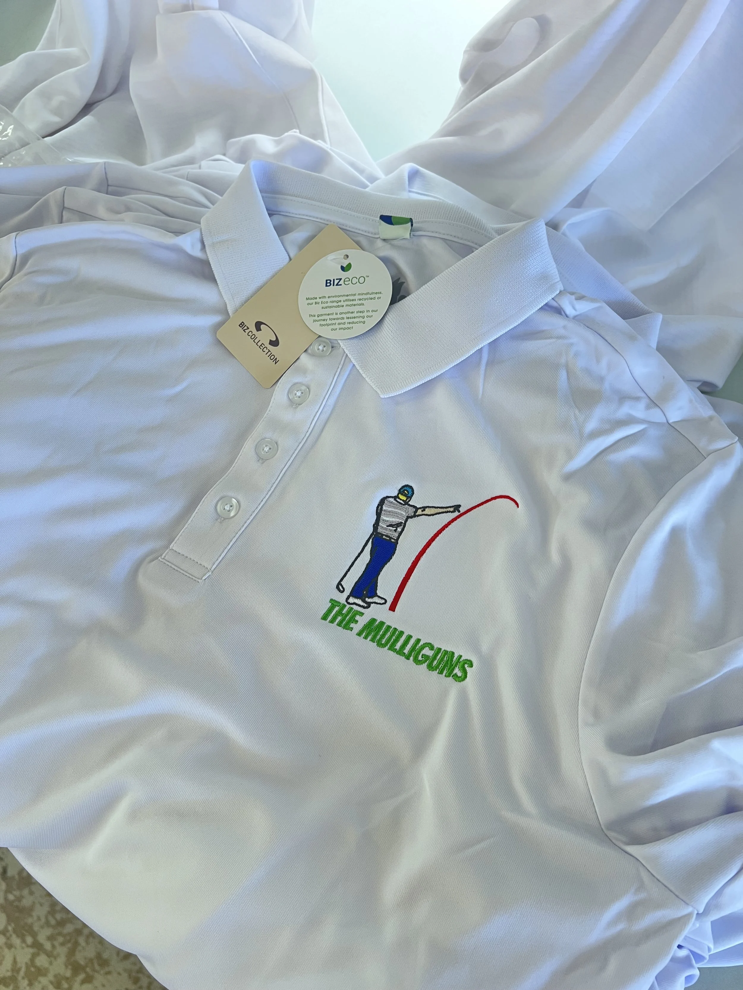 A white polo shirt with an embroidered logo of a golfer and text 'THE MULLIGUNS' on it. The shirt is laying on a white bedsheet.