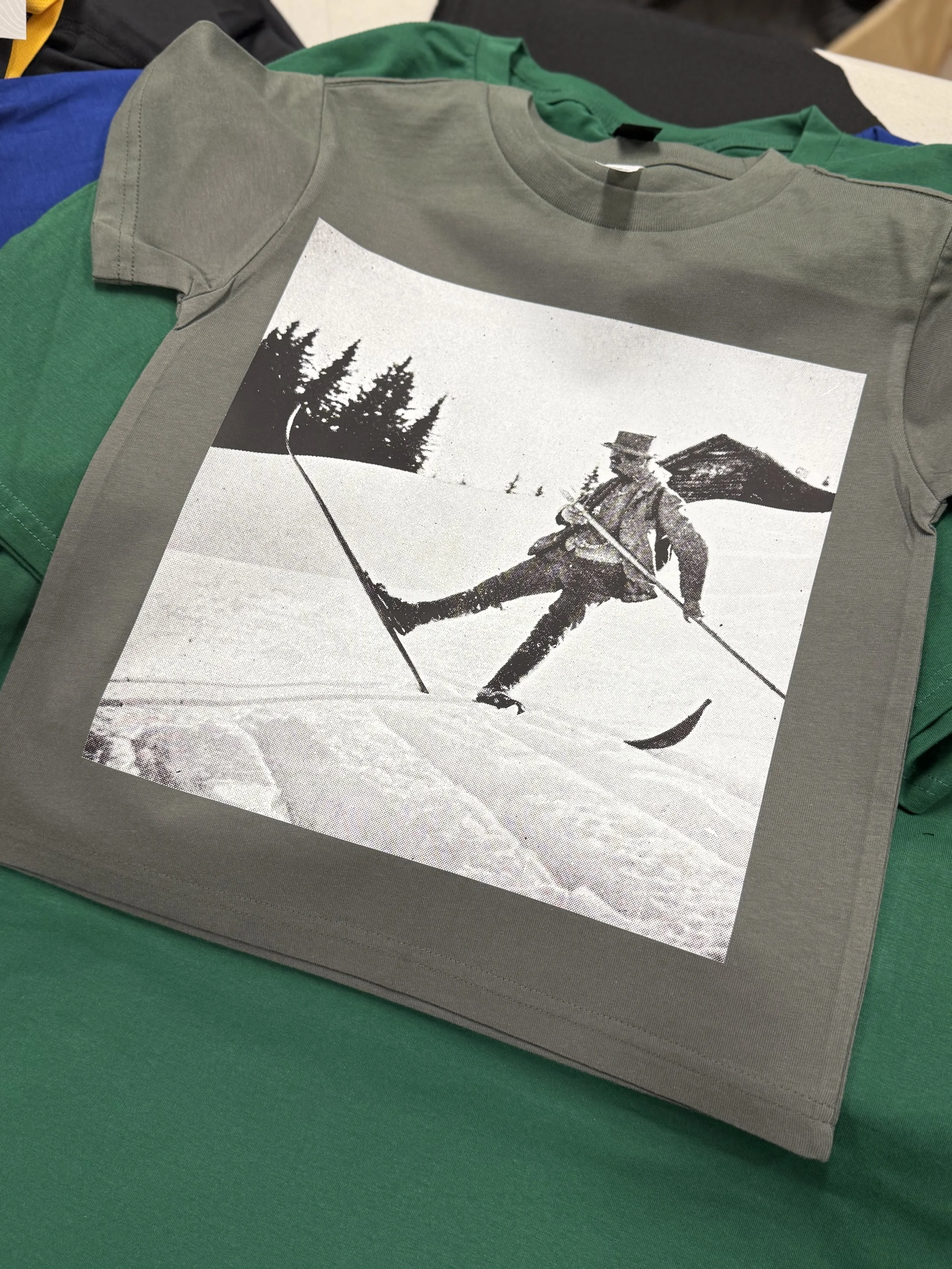 Gray T-shirt with a black-and-white photo of a person skiing in the snow near evergreen trees and a mountain in the background.