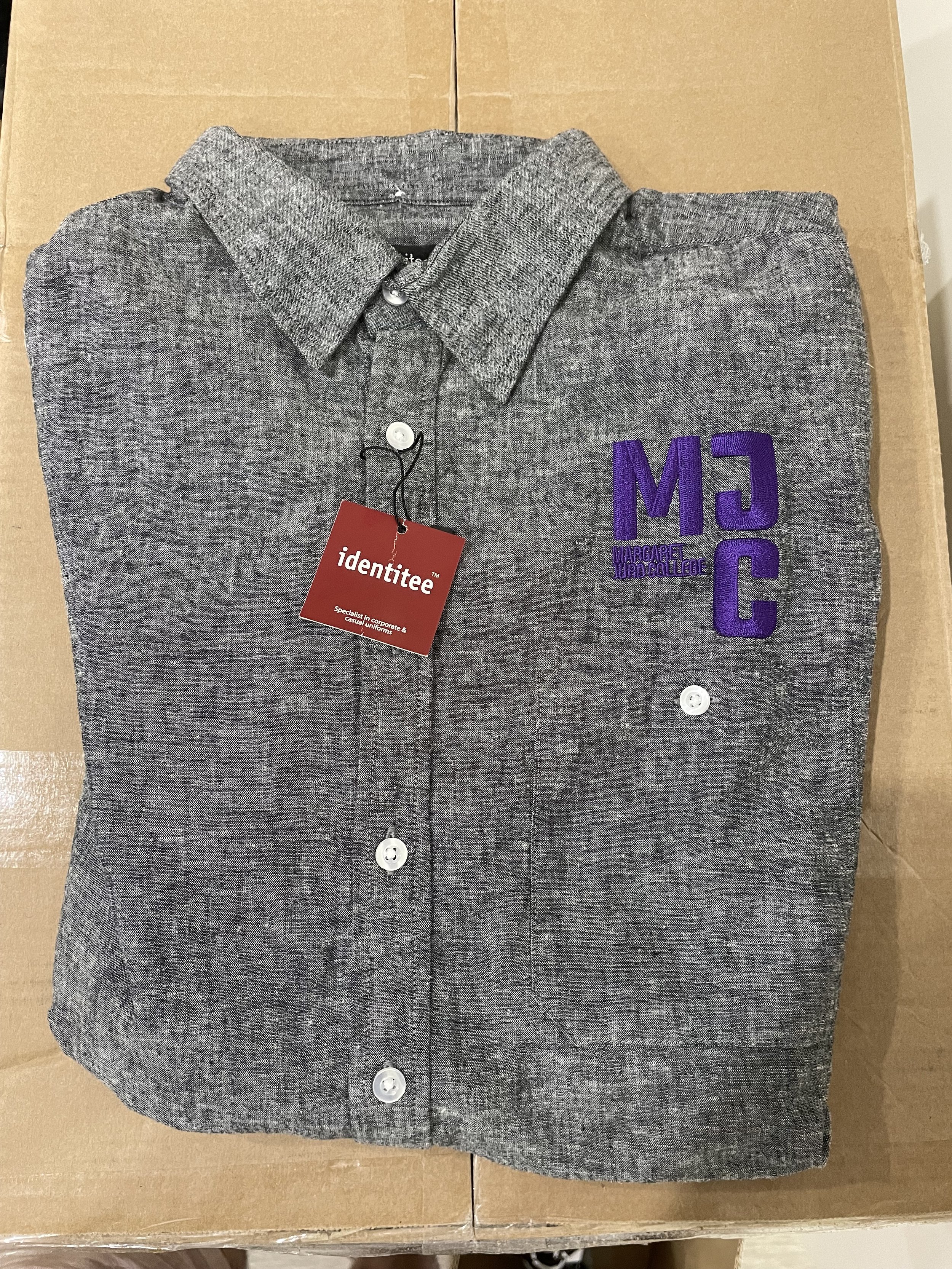 Folded gray collared shirt with purple embroidered letters 'MJC' on the left chest and a red tag that says 'identitee'.
