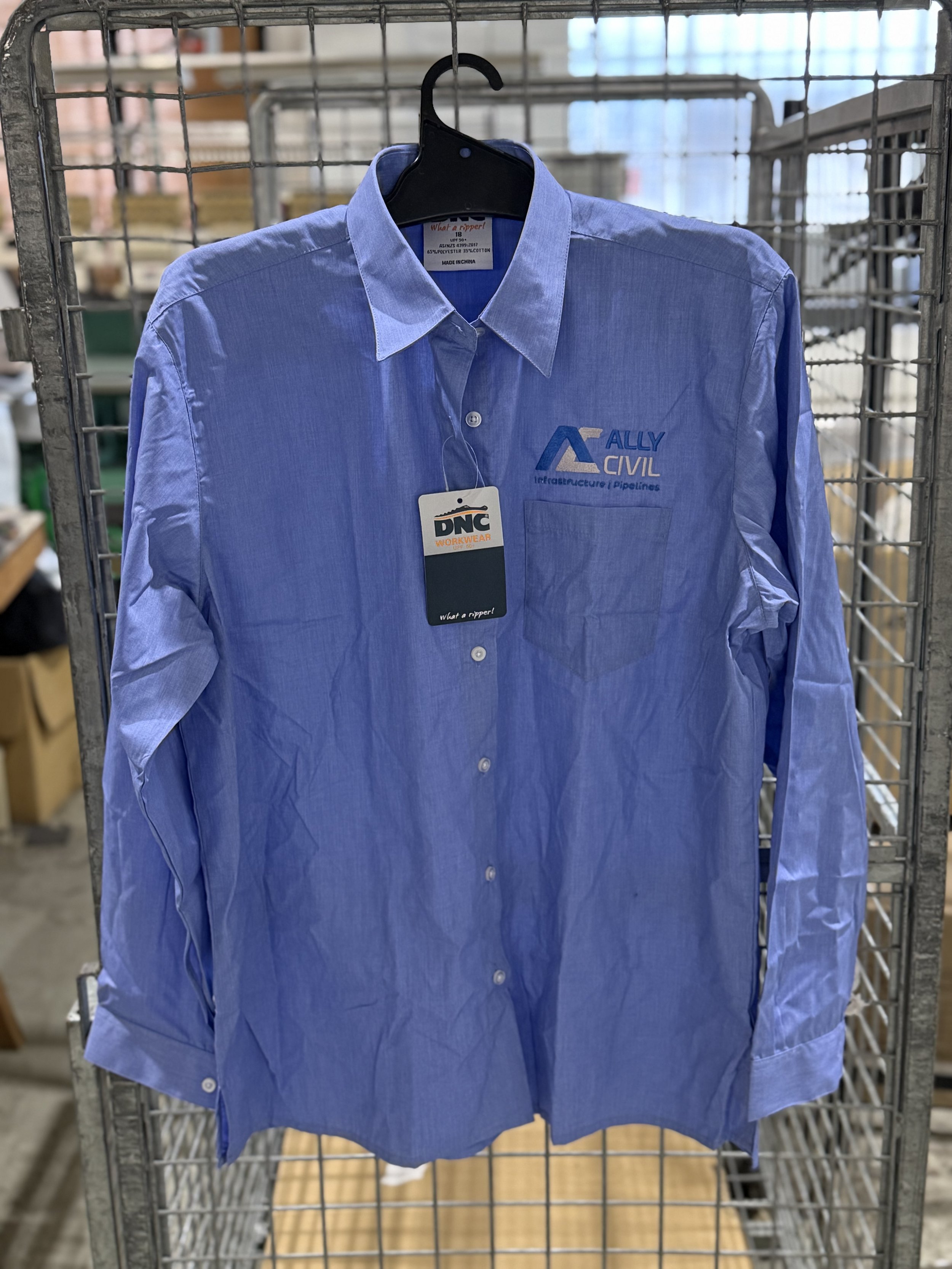 Blue work shirt on a hanger with logos for Ally Civil and DNC Workwear, hanging on a metal grid display.