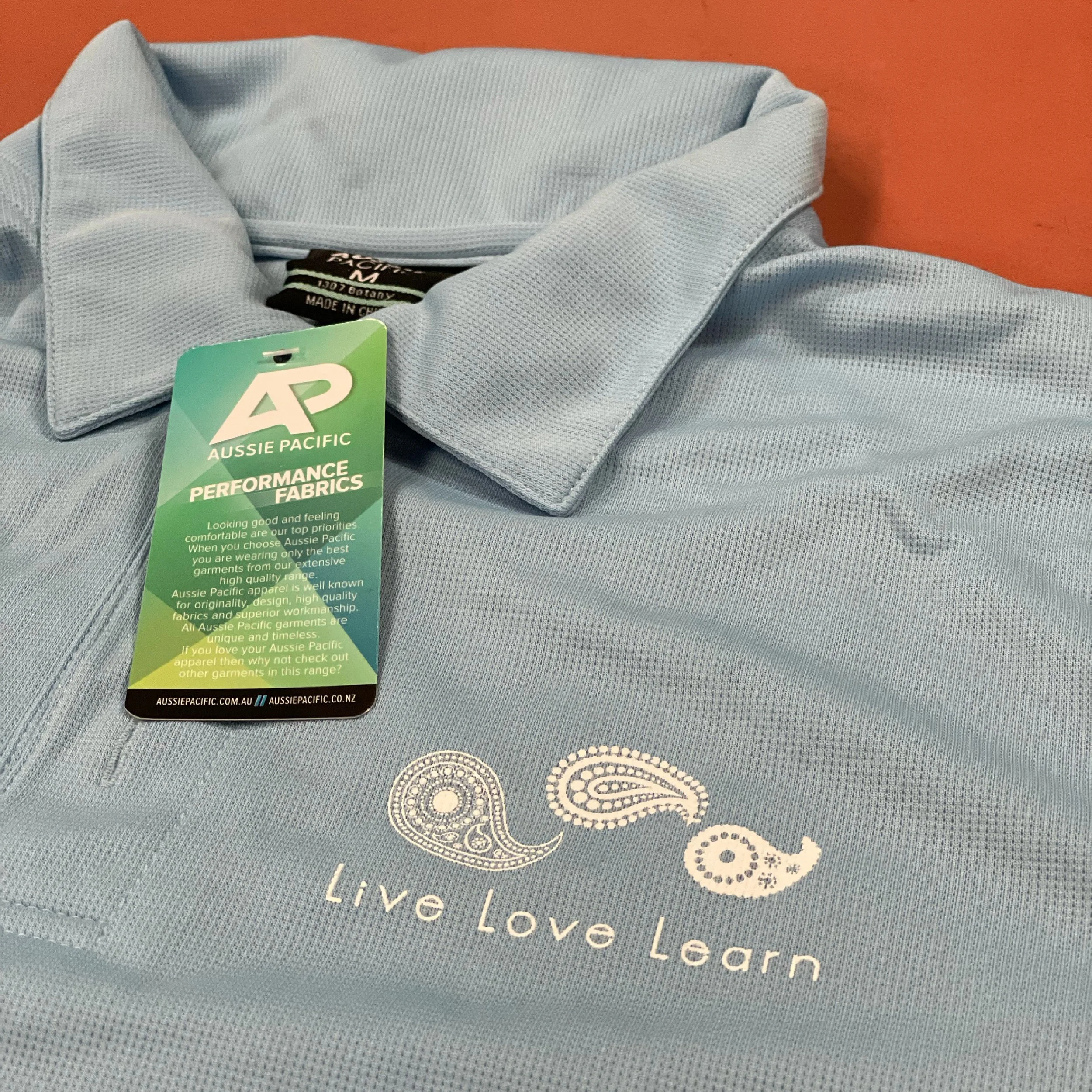 Light blue collared performance polo shirt with an 'Aussie Pacific' tag and a logo that says 'Eat, Live, Love, Learn' in white text on the front.