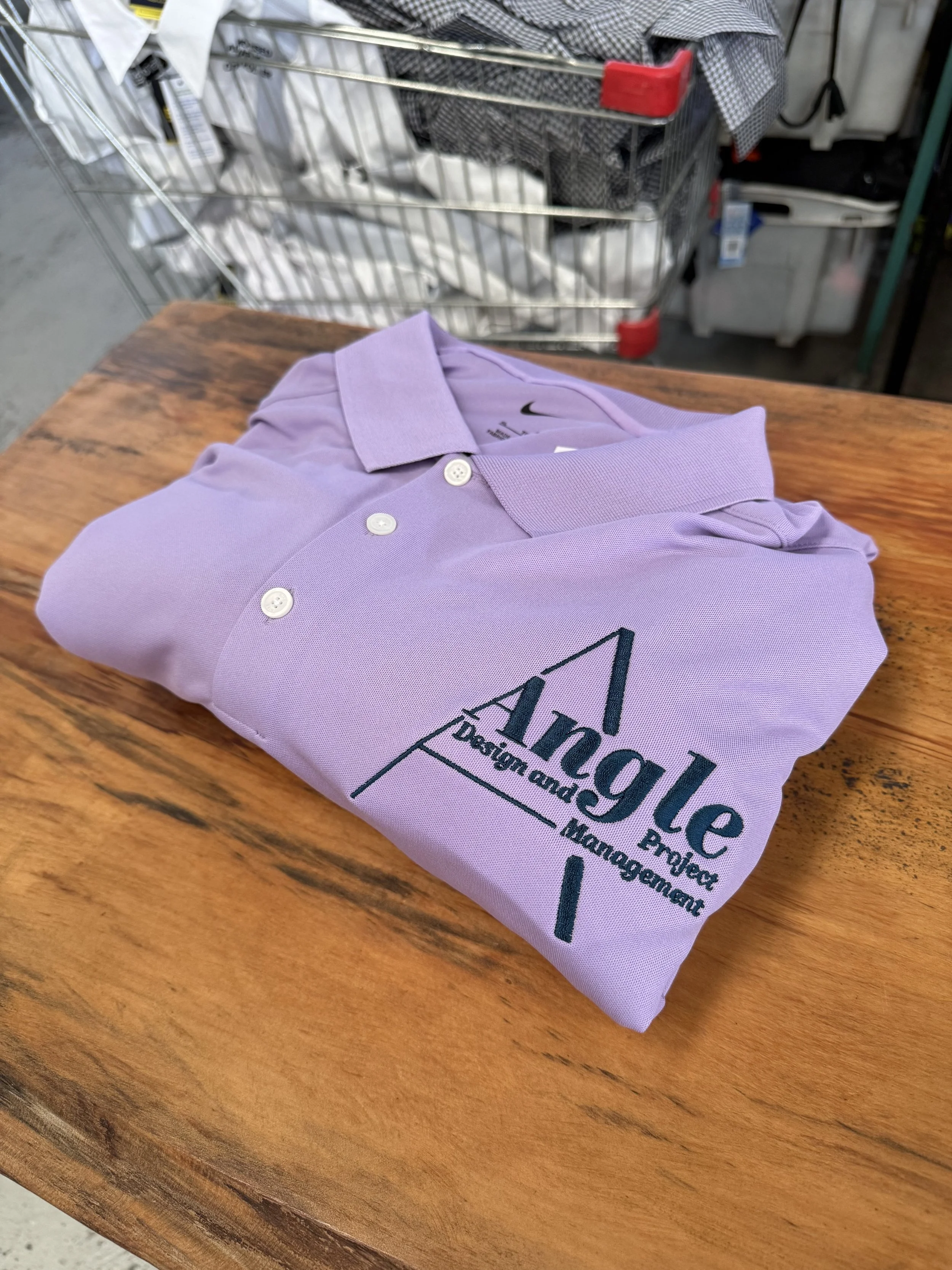 A folded lavender polo shirt with a logo that reads 'Angle Design and Project Management,' placed on a wooden surface with a shopping cart filled with various items in the background.