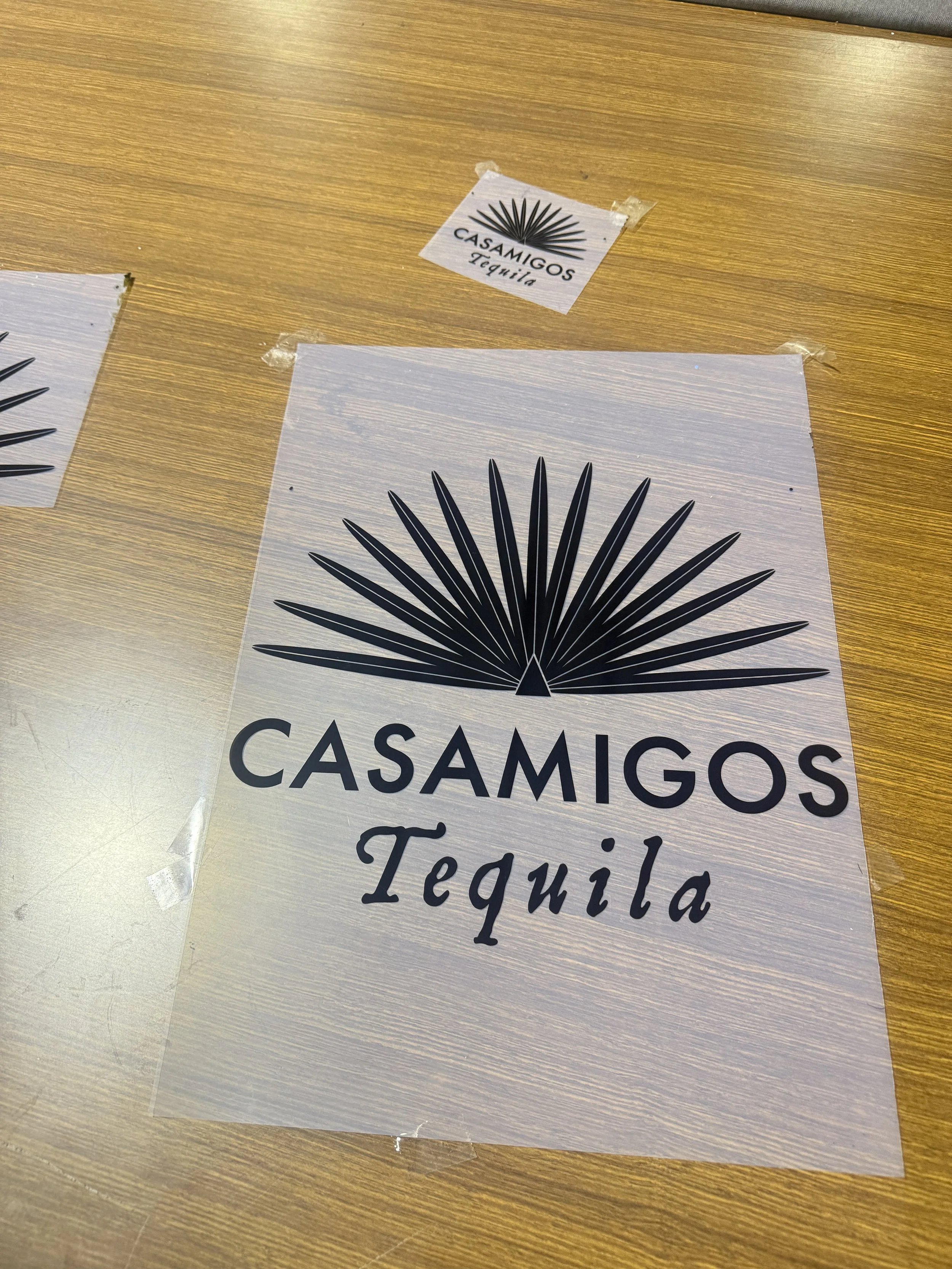 A printed sign for Casamigos Tequila with the brand's logo, featuring a stylized agave plant, on a wooden table.