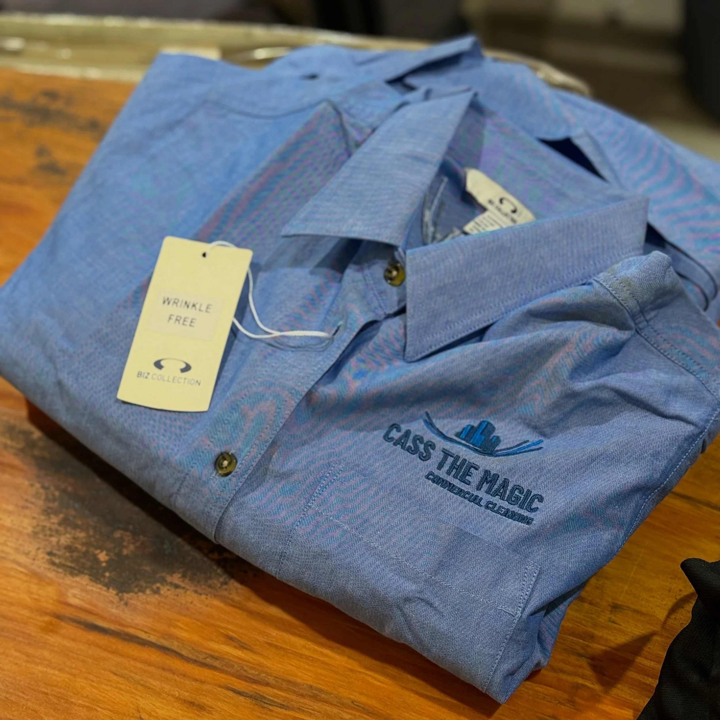 A folded blue collared shirt with embroidered logo for 'Class The Magic Commercial Cleaning' on a wooden surface, with a tag indicating 'Wrinkle Free'.