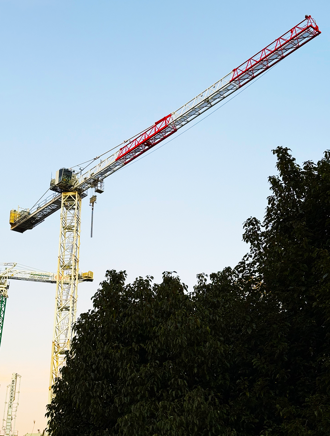 A construction crane with a yellow base, long white and red arm, set against a clear blue sky, with green trees at the bottom of the image.