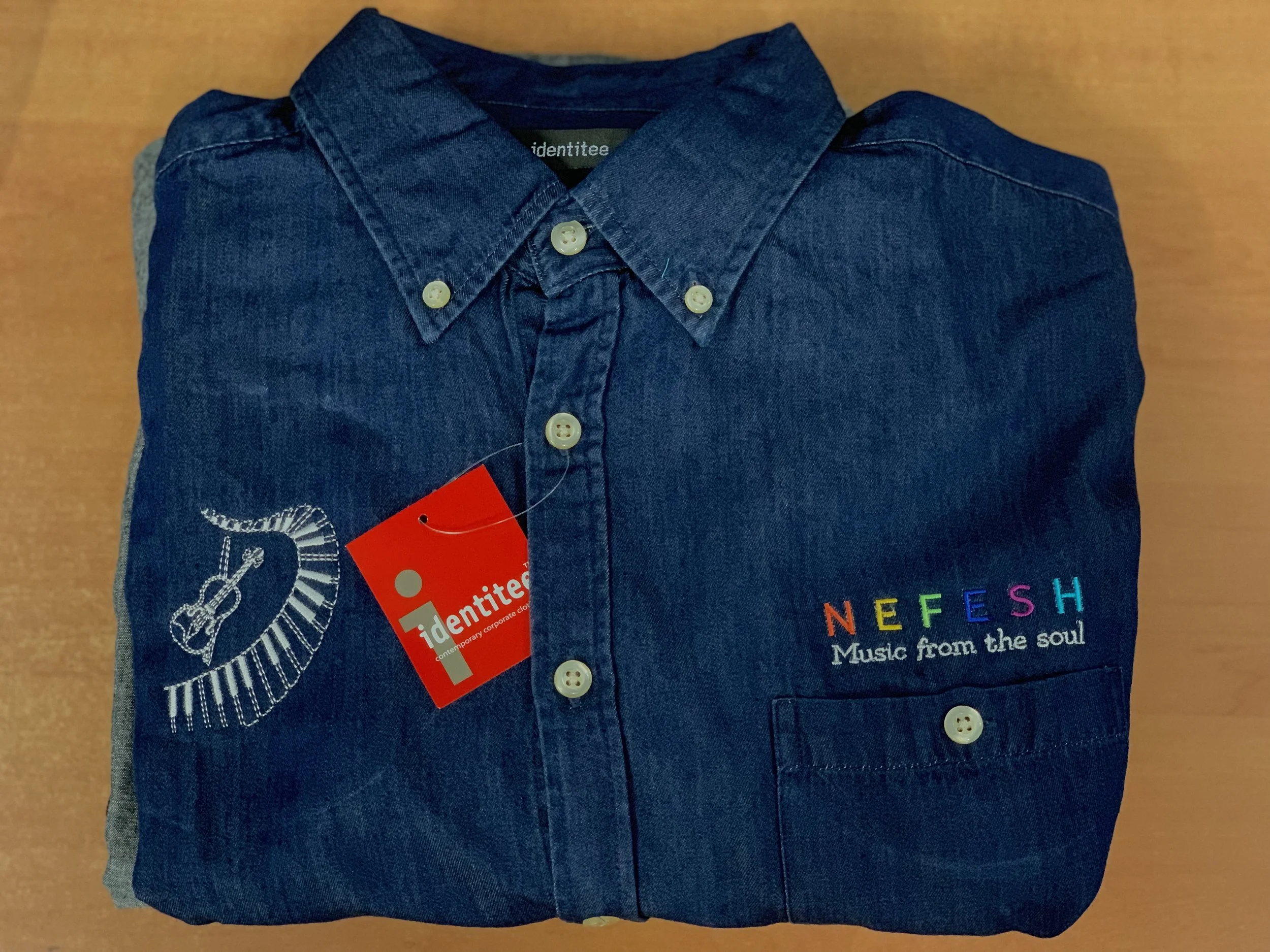 Folded denim shirt with embroidered guitar and colorful 'NEFESH' text, sitting on a wooden surface