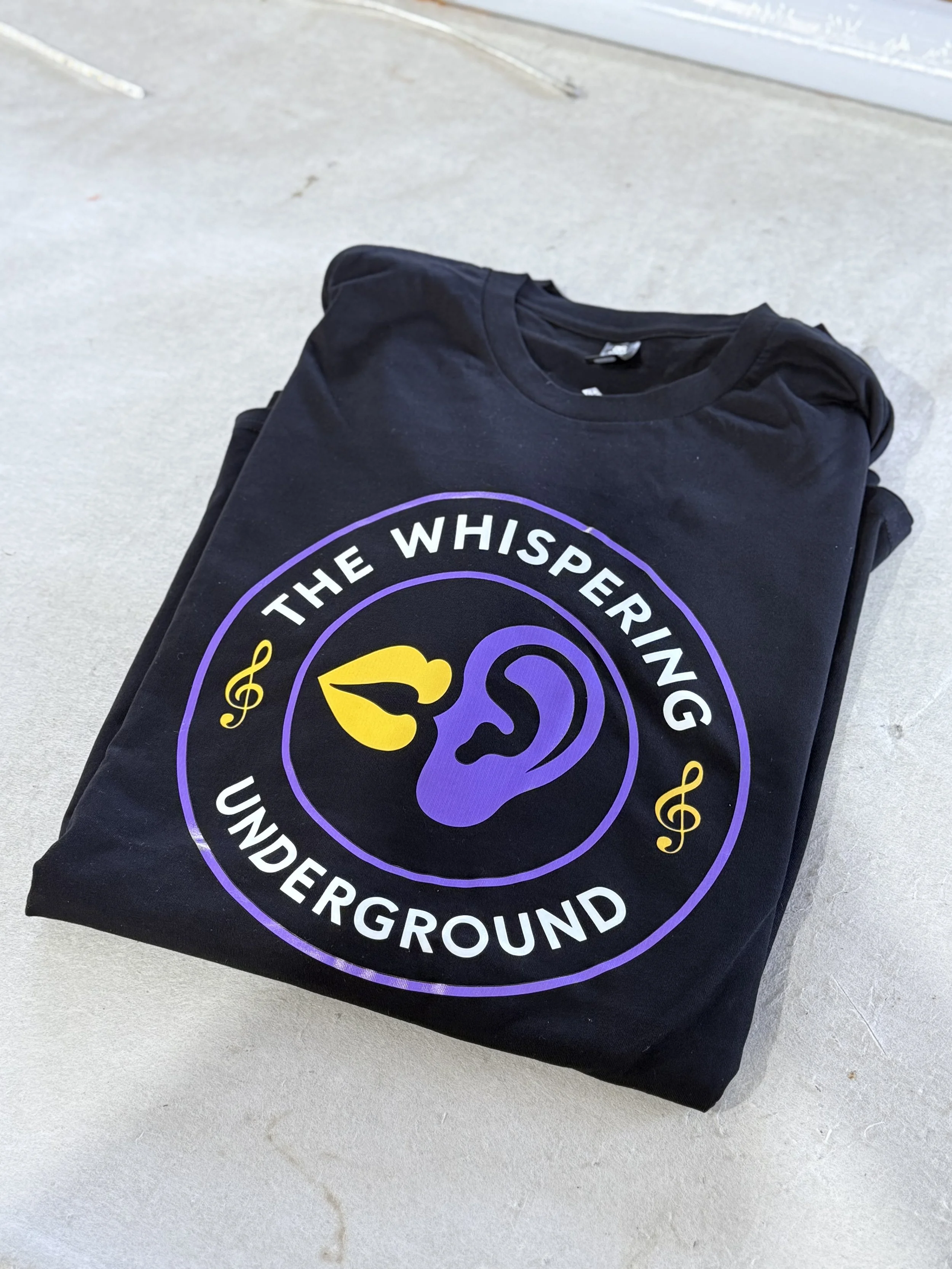 Black t-shirt with a circular logo reading 'The Whispering Underground,' featuring a purple ear, yellow lips, and yellow musical notes, laid on a concrete surface.