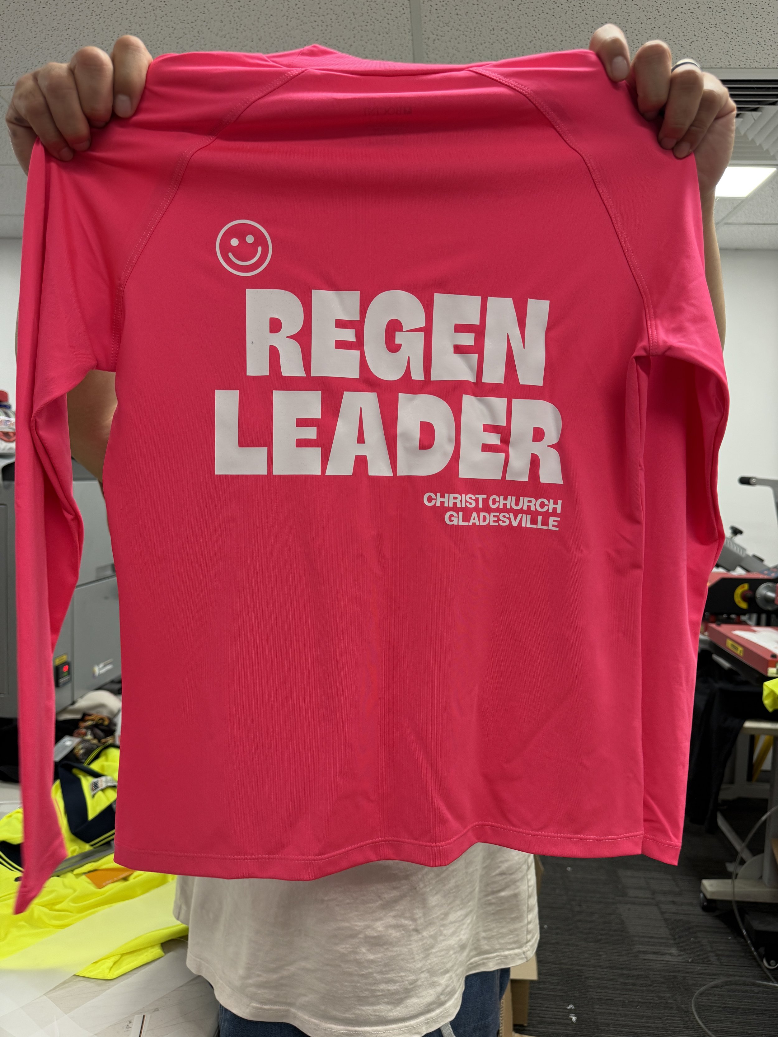 A person holding a bright pink shirt with white text and a smiley face. The shirt reads 'REGEN LEADER' with smaller text below that says 'CHRIST CHURCH GLADSVILLE'.