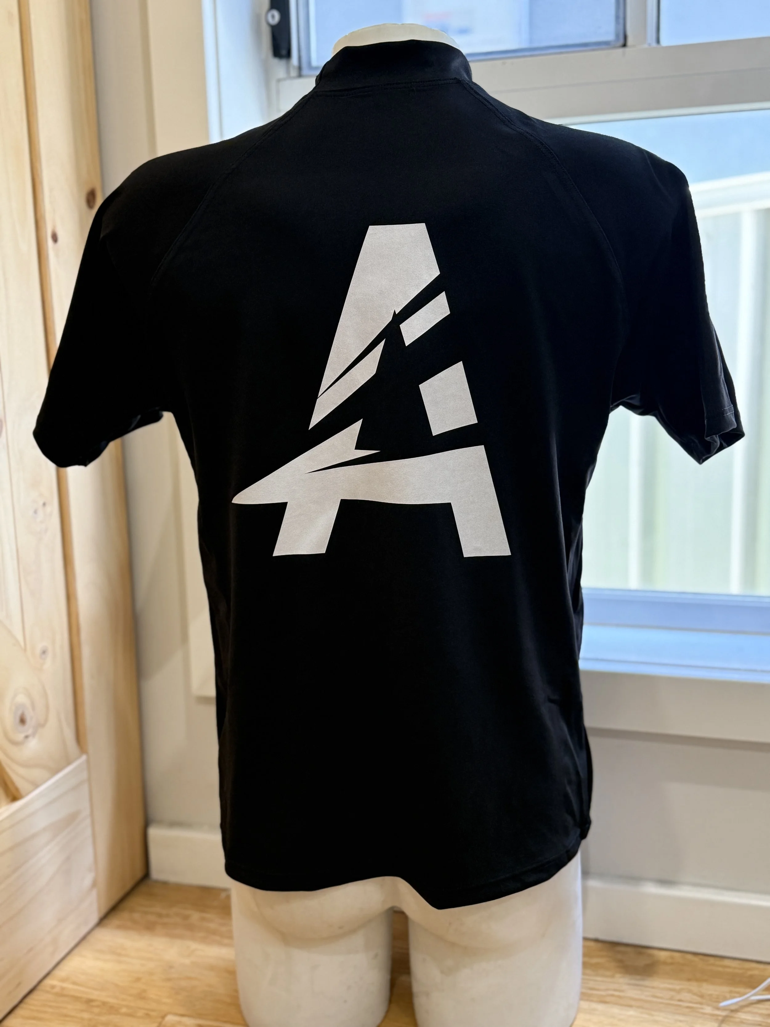 Black T-shirt with a white graphic logo on the back, featuring a stylized letter 'A' with a lightning bolt design, displayed on a mannequin torso near a window.