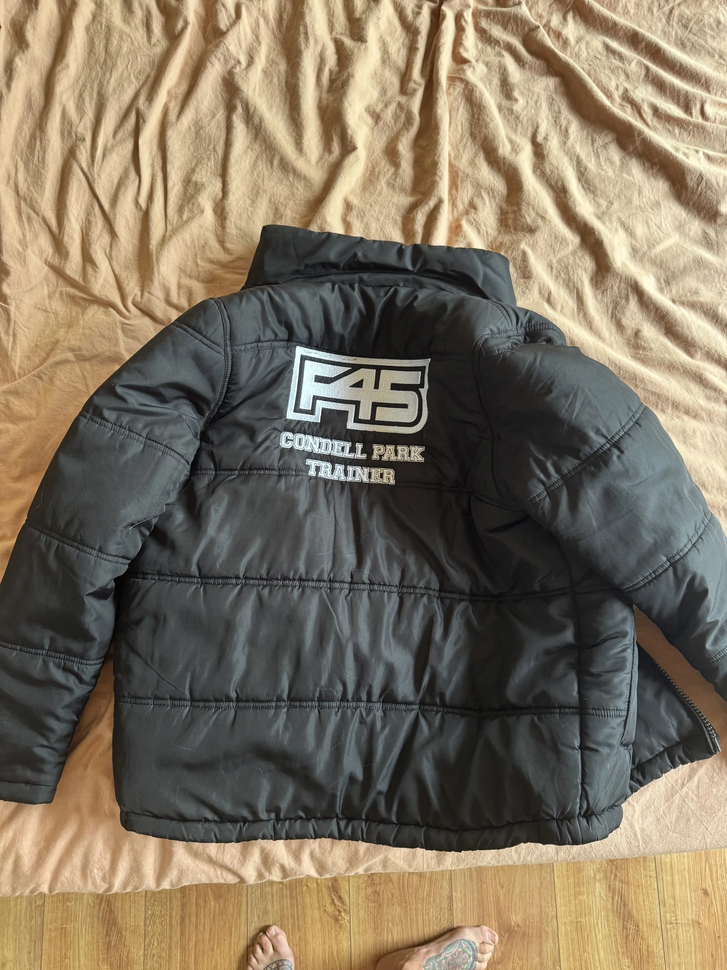 Black padded jacket with embroidery reading 'F4S', 'Condell Park', and 'Trainer' laid on a beige bedsheet.