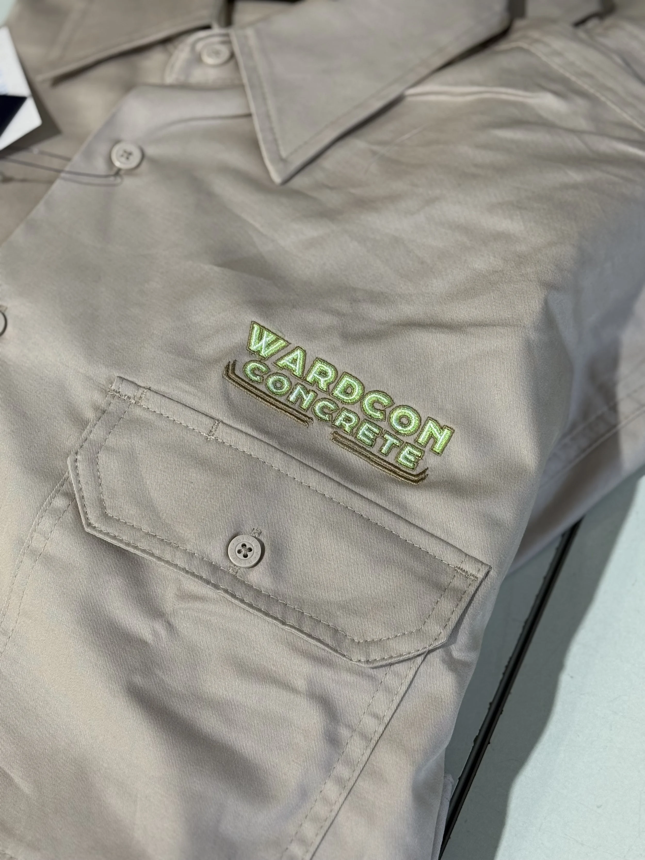 Close-up of a beige shirt with an embroidered logo that says 'WARDCON CONCRETE' in green and gold, with a buttoned chest pocket.