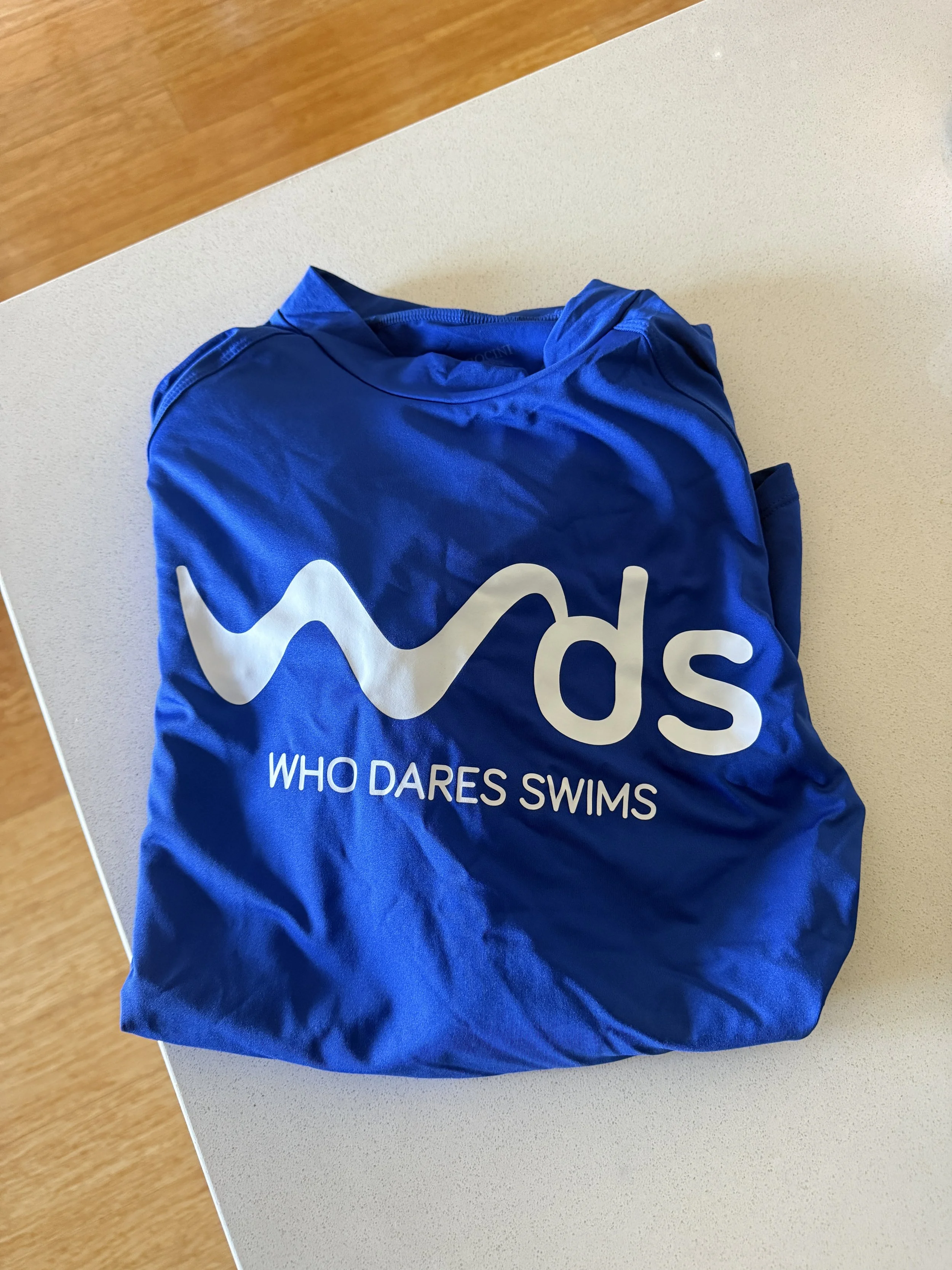 A blue swimsuit with the words 'WHO DARES SWIMS' printed on it, folded on a beige table.