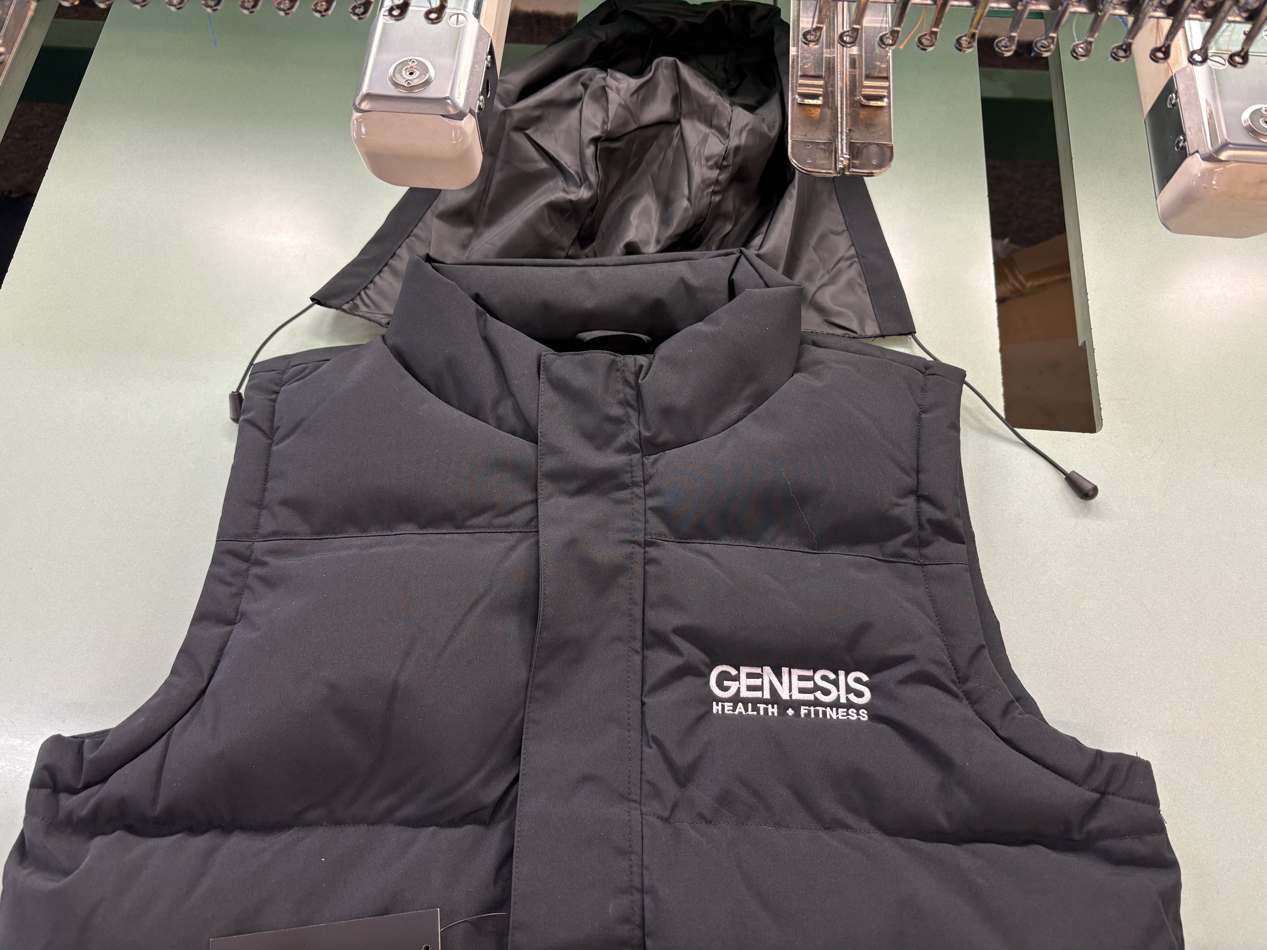 Black puffer vest with 'GENESIS HEALTH + FITNESS' logo on the front, laid out on a table with sewing and embroidery equipment in the background.