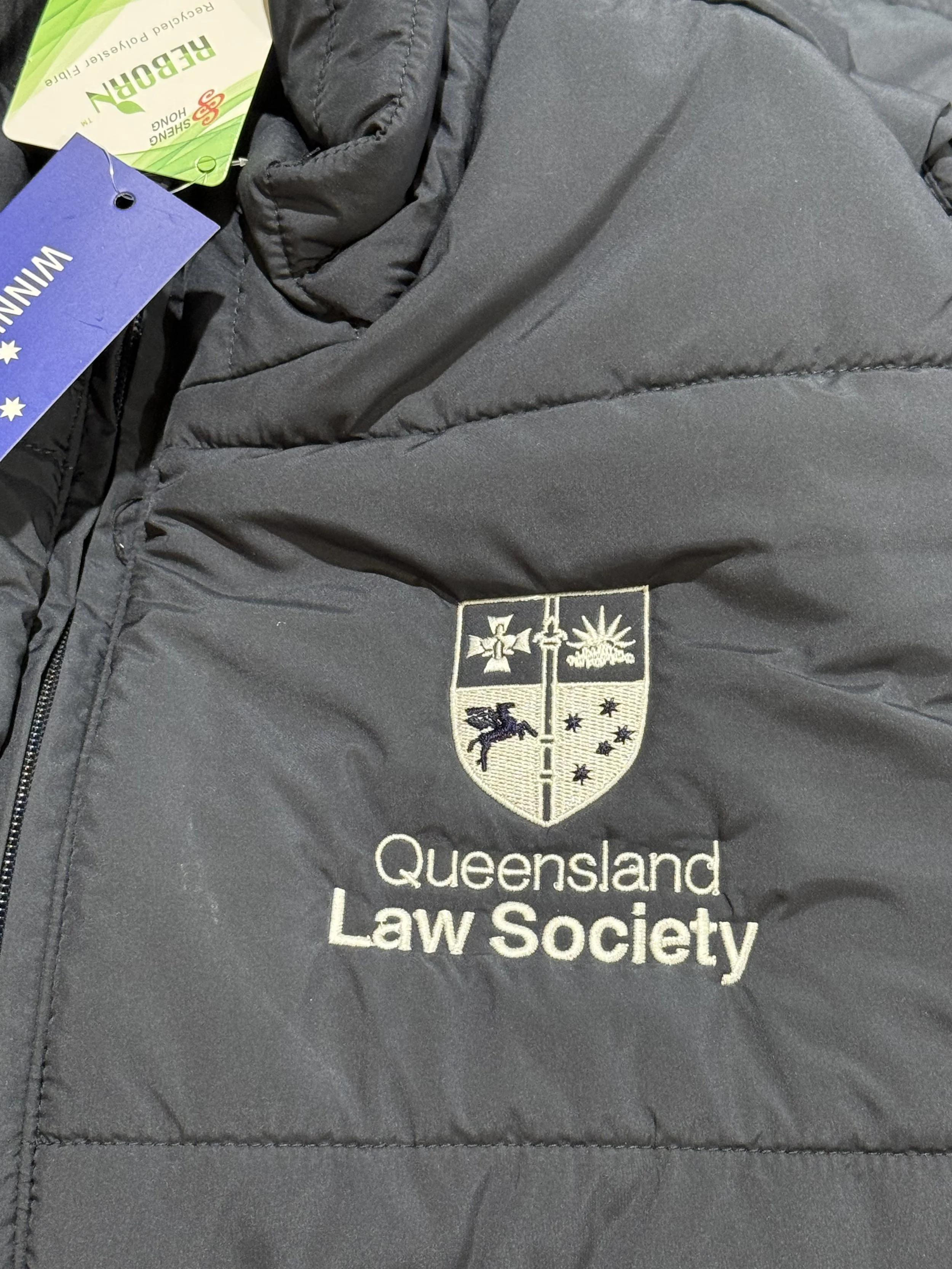 A gray quilted jacket with an embroidered patch that reads "Queensland Law Society" and features a shield with symbols including a cross, a star, and a mythical creature.
