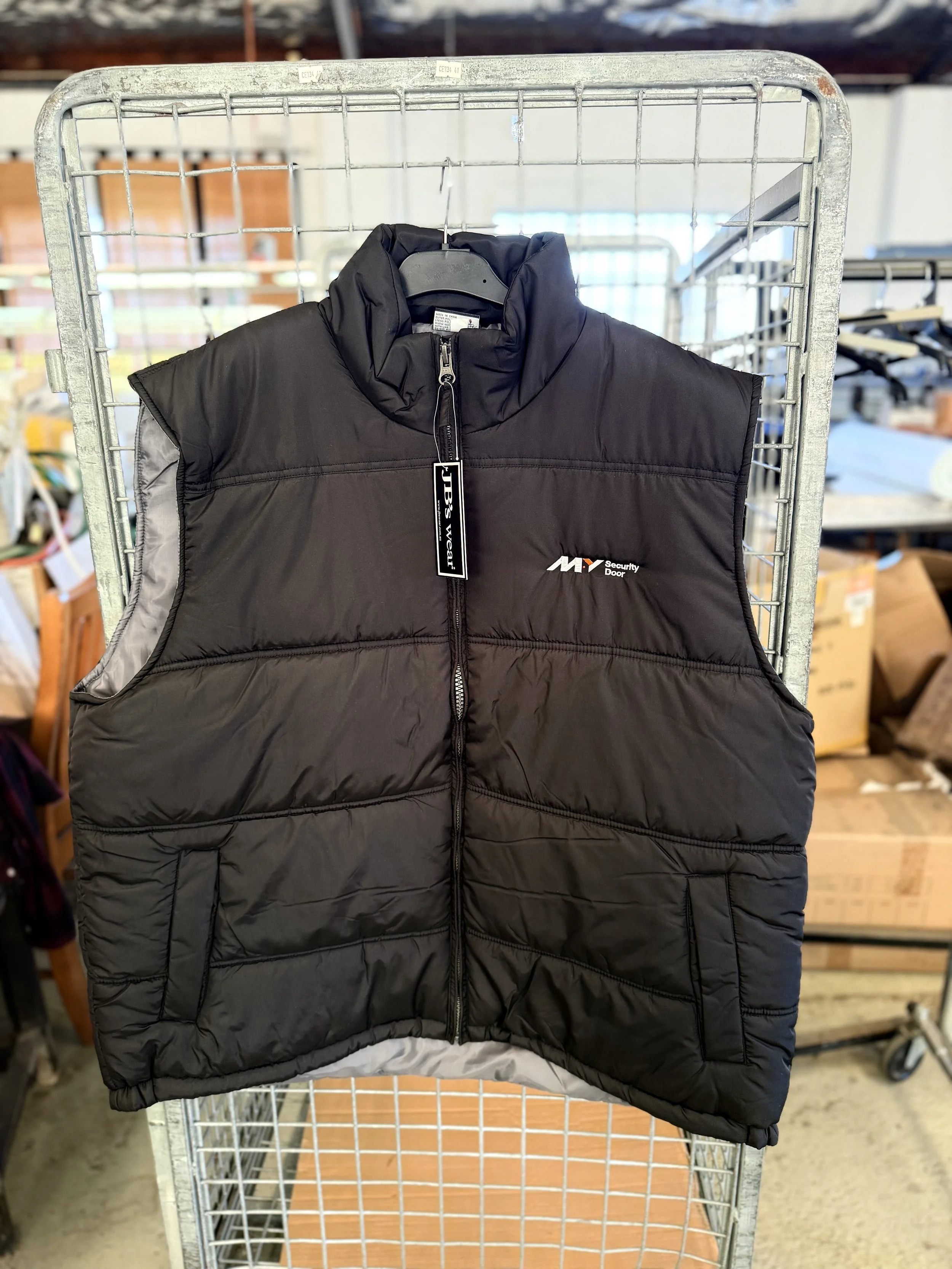 Black puffy sleeveless vest with a full front zipper and a logo on the chest hanging on a hanger inside a retail store.