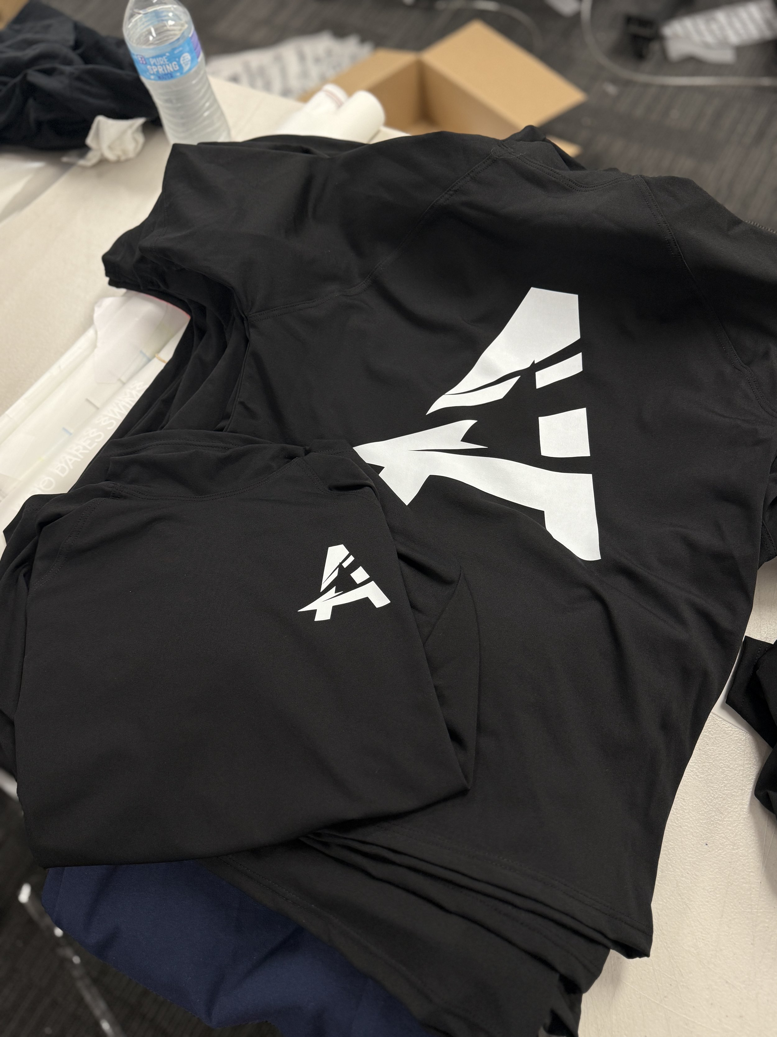 Black T-shirts with white logo on a table, one shirt folded and one laid out, near a water bottle and other items.