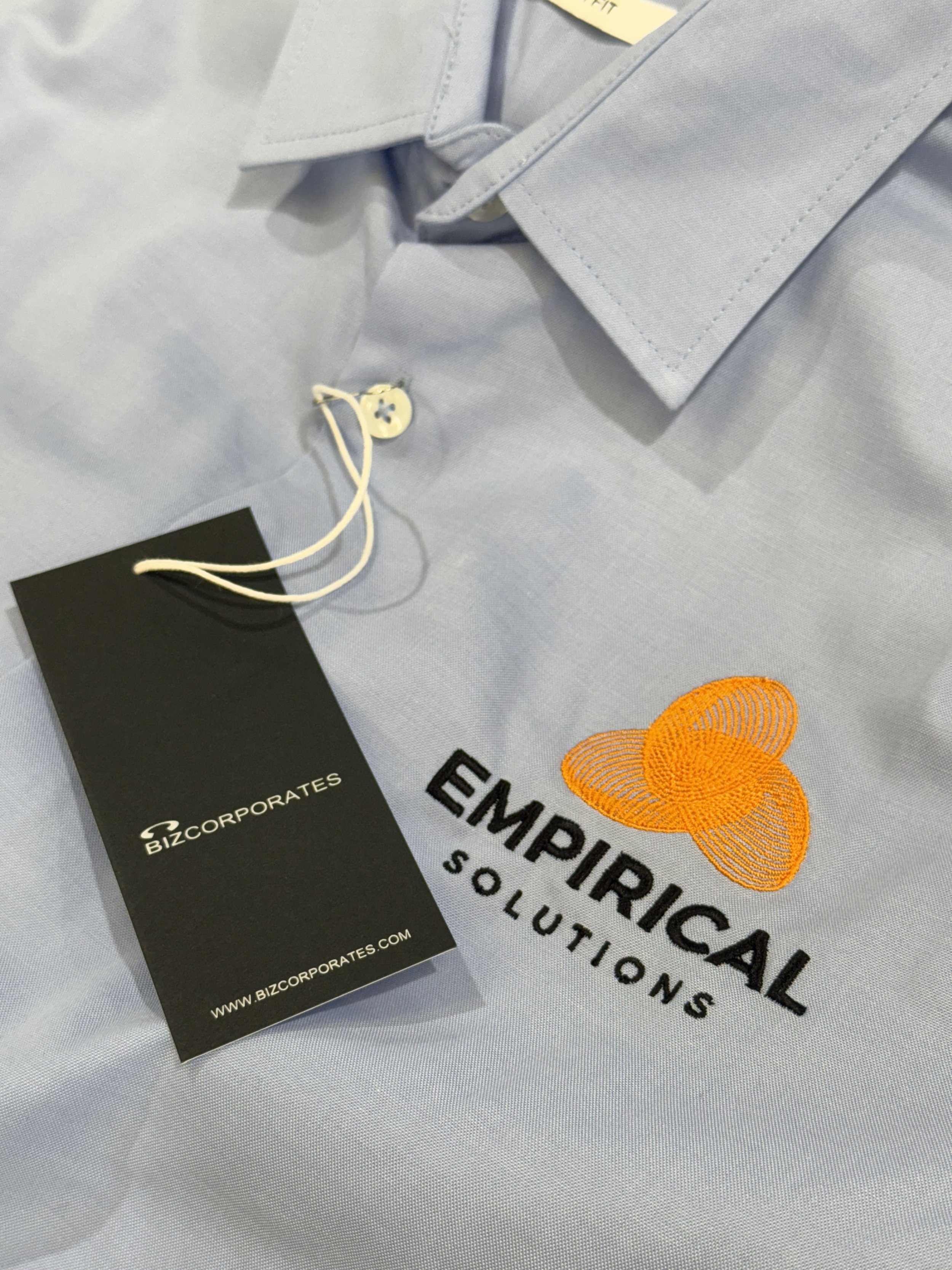 Light gray shirt with embroidered logo of three orange interconnected circles and the words 'EMPIRICAL SOLUTIONS' underneath, along with a black tag showing 'BizCorporates' branding.