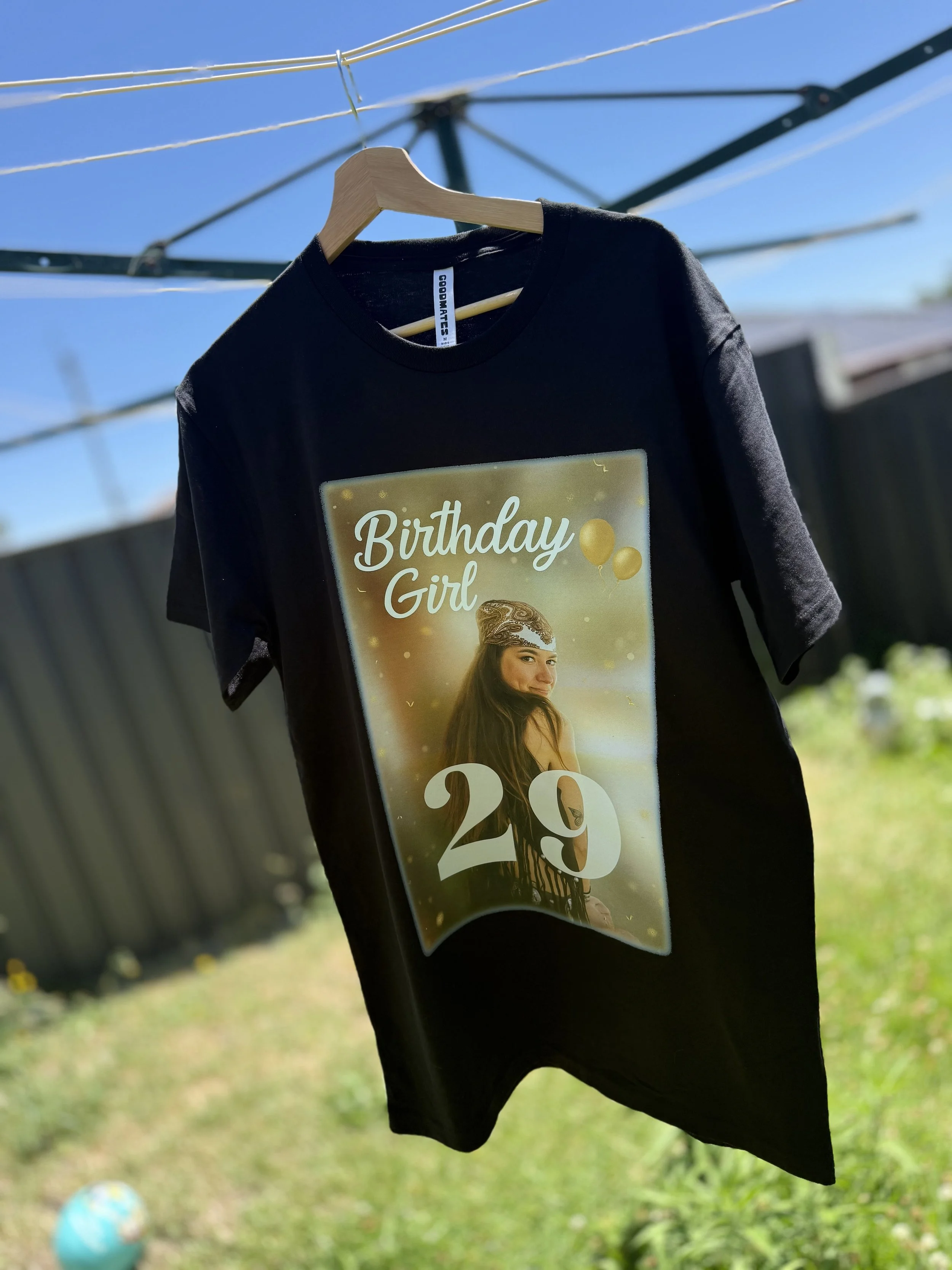 A black T-shirt hanging on a wooden hanger outdoors, featuring a birthday design with a girl, balloons, and the text 'Birthday Girl 29'.