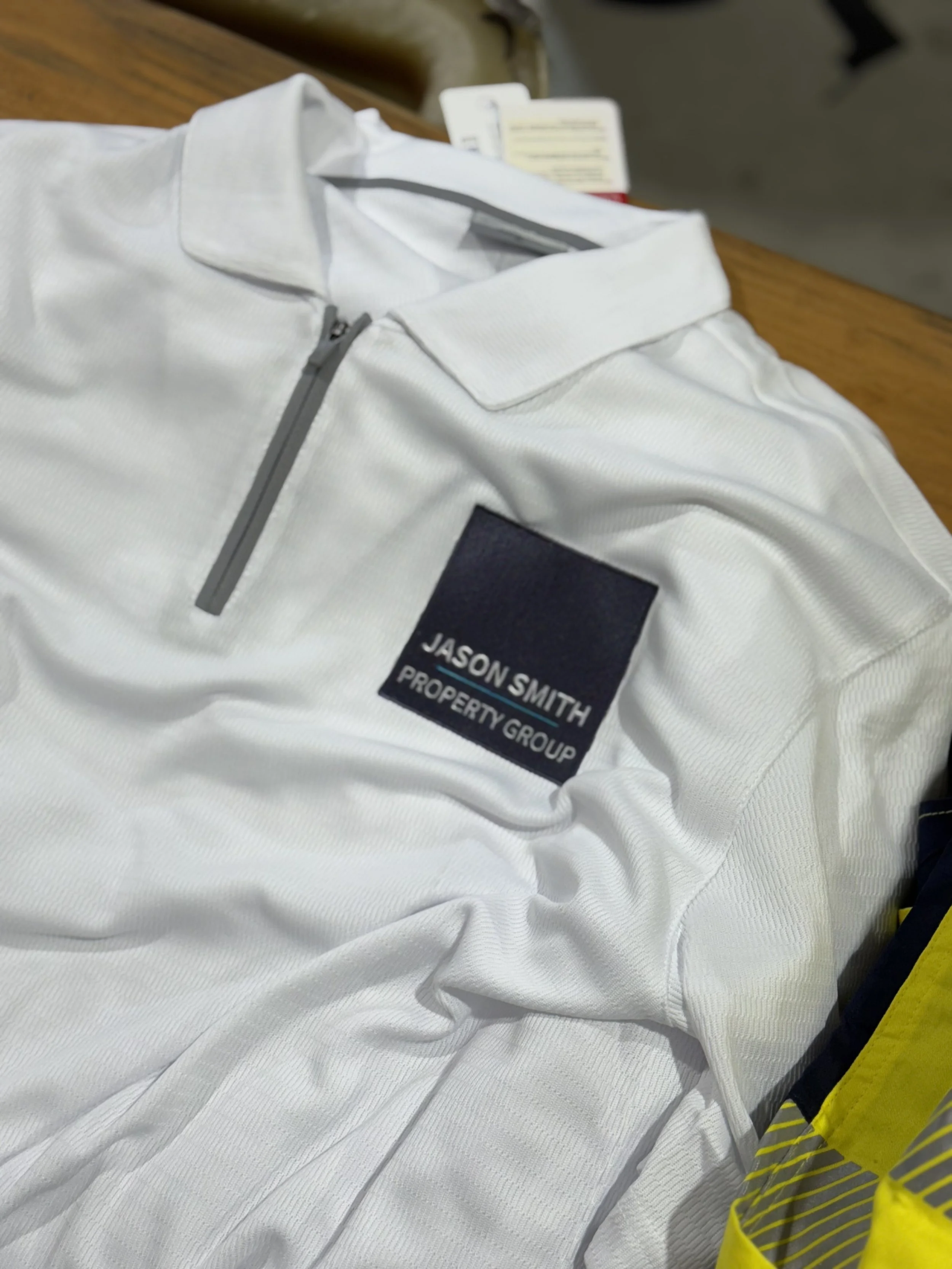 White polo shirt with a zippered collar and a navy patch that reads 'Jason Smith Property Group' lying on a wooden surface.