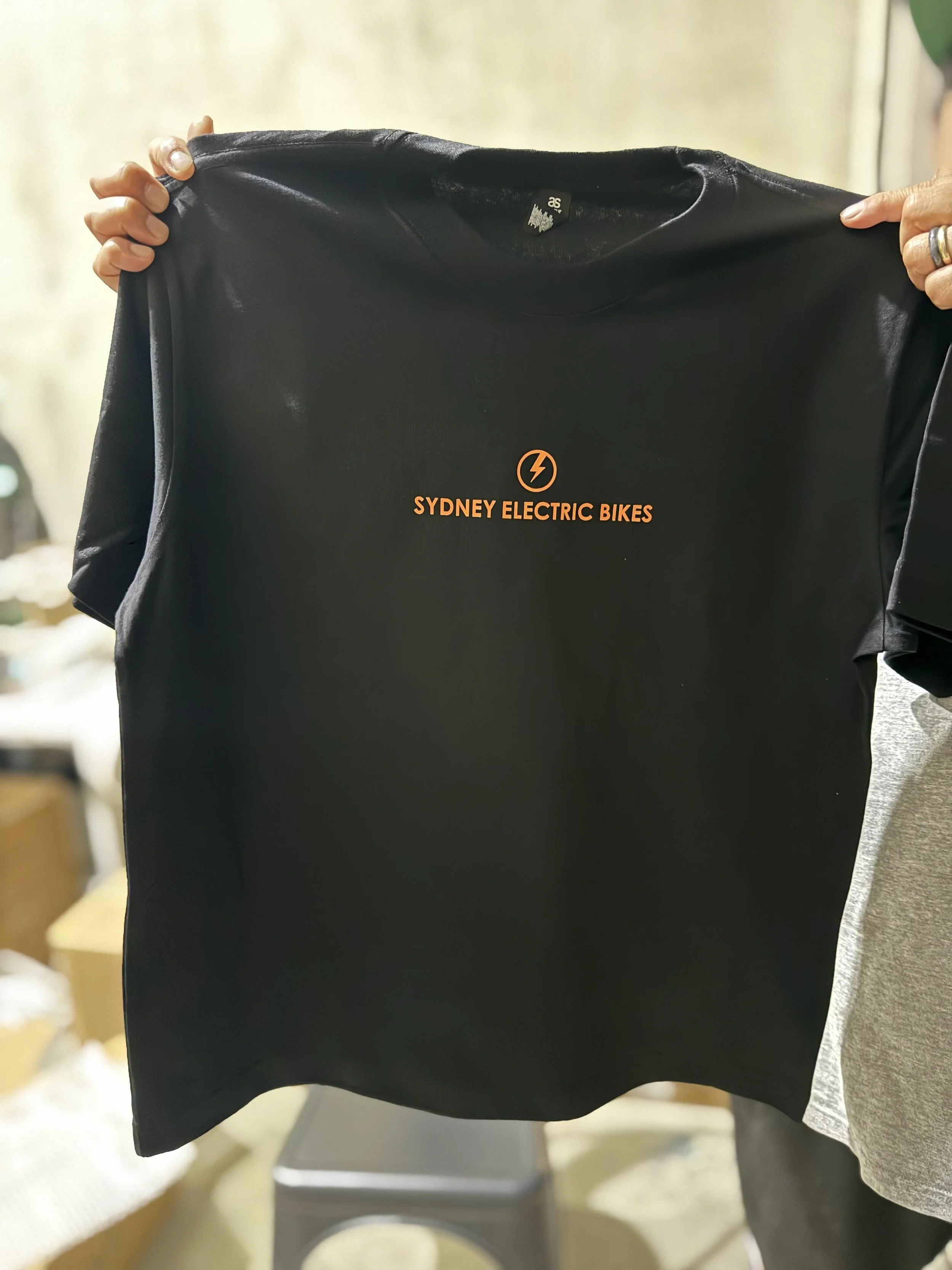 Black T-shirt with orange text and logo reading "Sydney Electric Bikes" being held up by two hands.