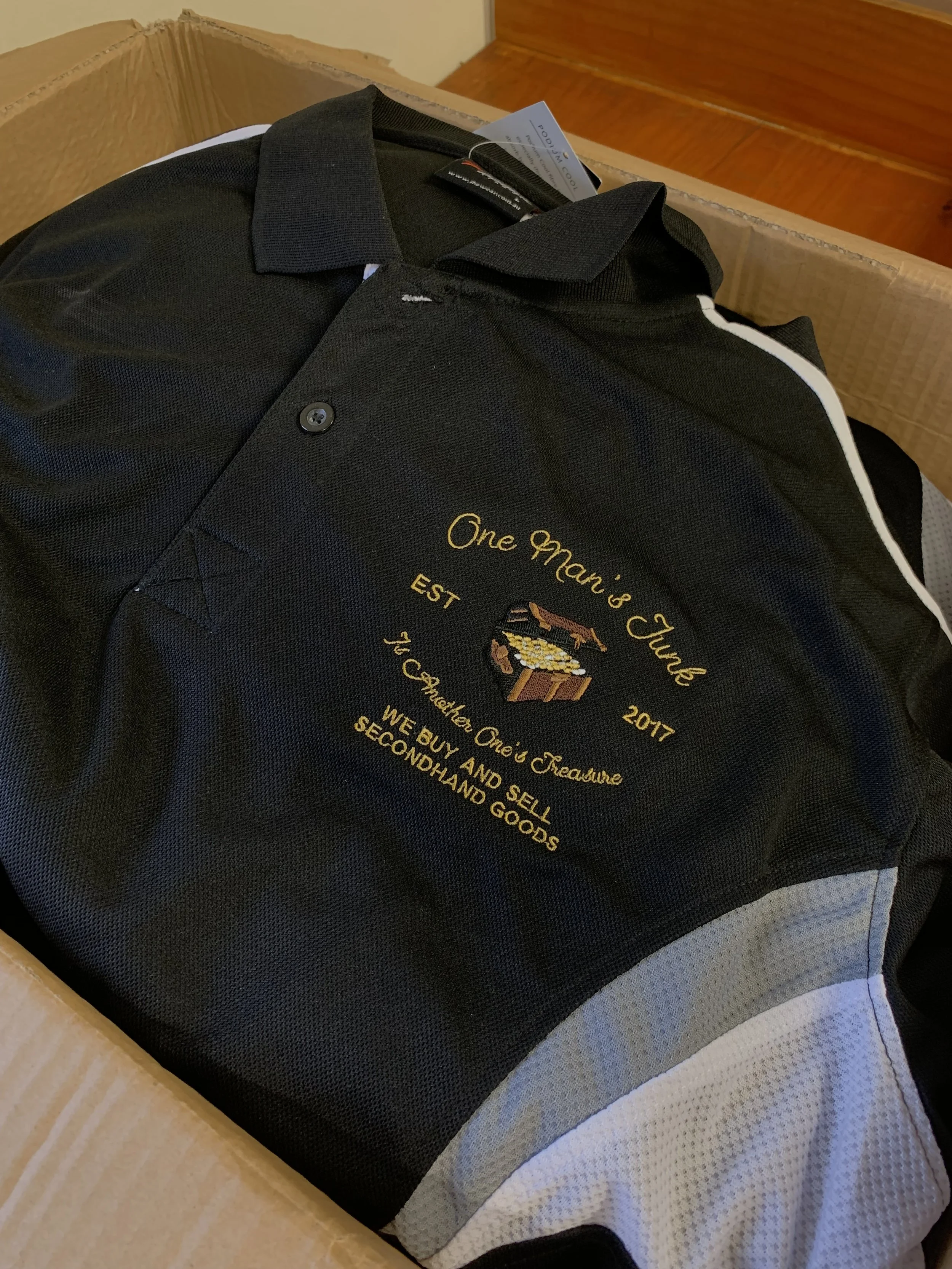 A black embroidered polo shirt inside a cardboard box. The shirt features embroidery with the text 'One Man's Junk,' 'EST 2017,' and 'Yard Sale' along with a treasure chest illustration and the phrase 'We buy and sell secondhand goods.'