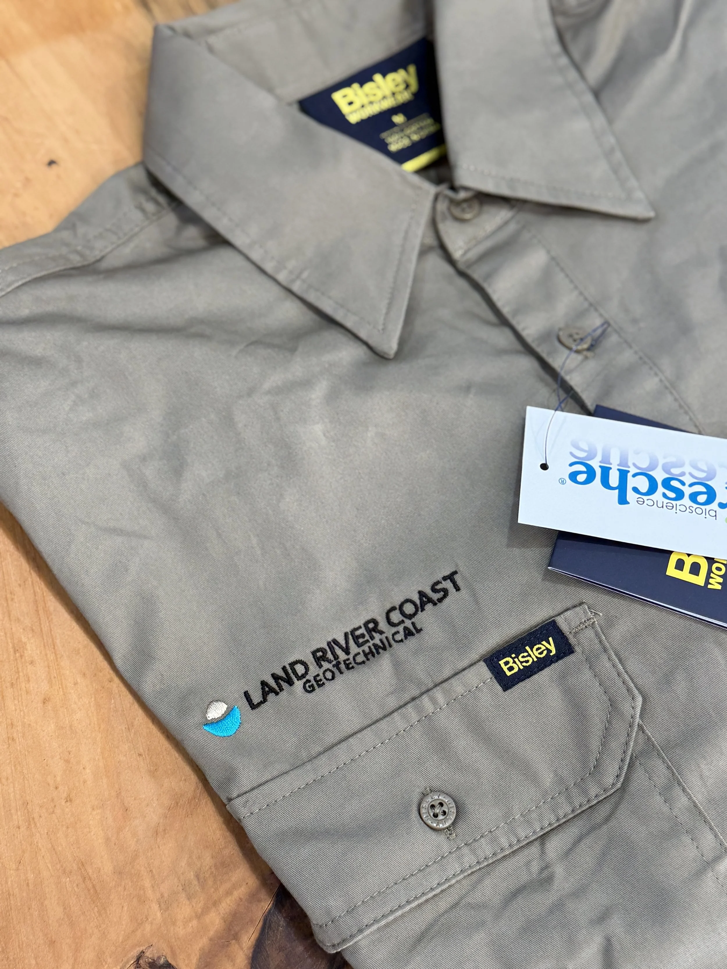 Gray long-sleeve work shirt with a collar, embroidered with the text "Land River Coast Geotechnical" and two logos, one depicting a wave and the other labeled "Bisley," placed on a wooden surface with a tag attached.