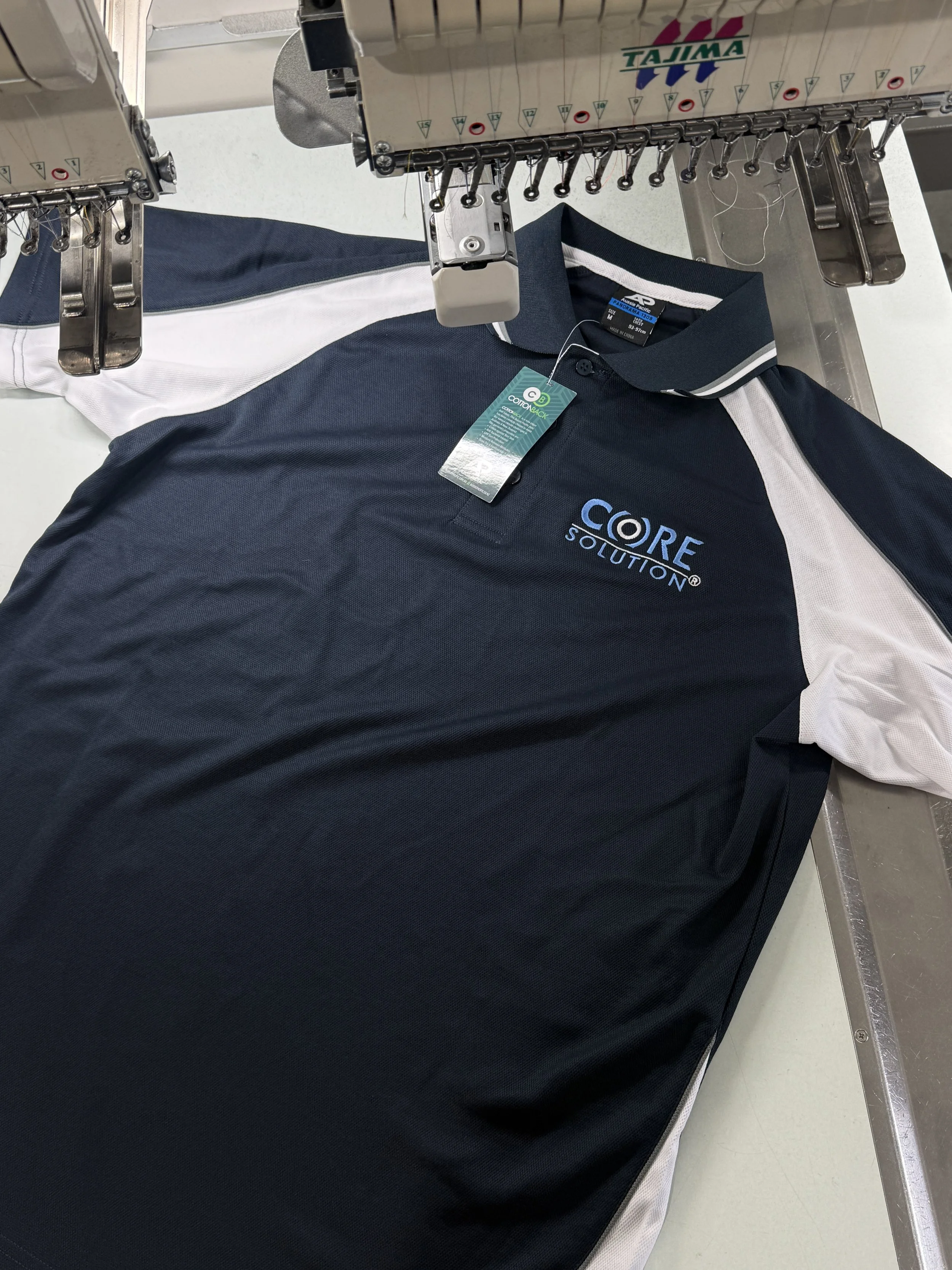 A navy blue and white polo shirt with the logo 'Core Solution' is on a machine used for embroidery or sewing.