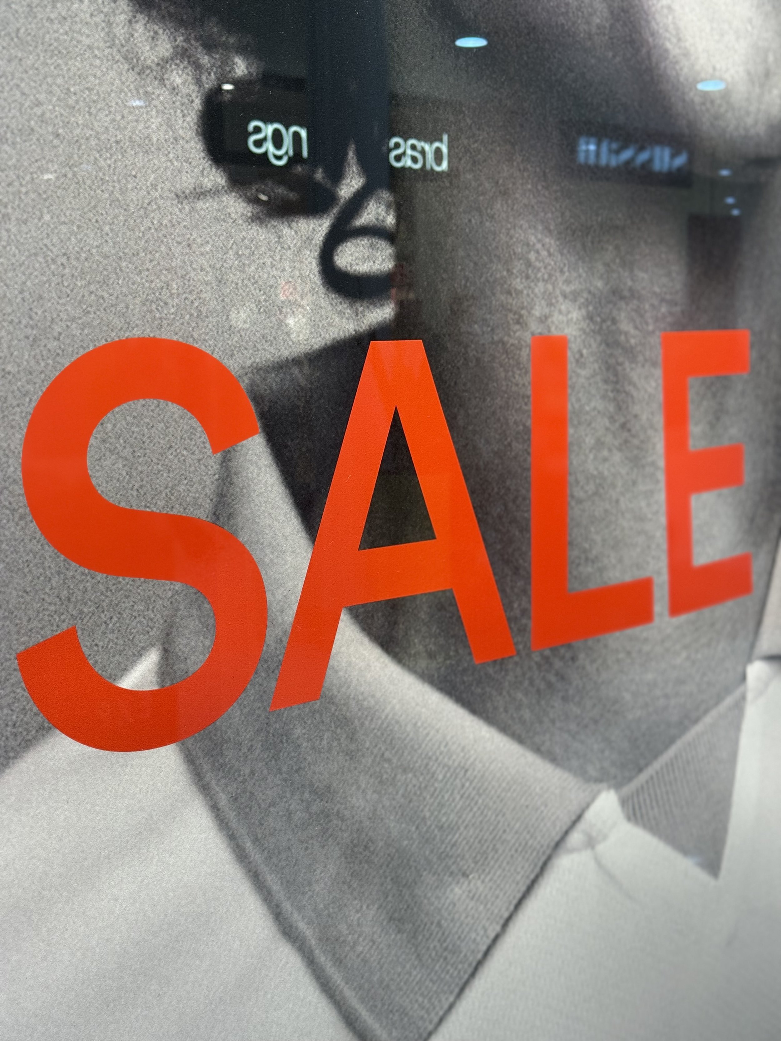 Glass window with red 'SALE' sign. Behind the glass, there is a reflection of a person wearing glasses and a shirt. There are also some blue and white illuminated signs in the background.