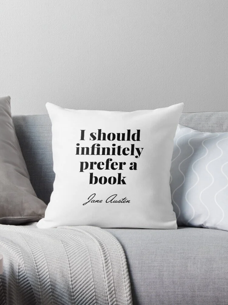 White pillow with black text that reads: 'I should infinitely prefer a book' by Jane Austen, on a light gray sofa with other pillows and a gray blanket.
