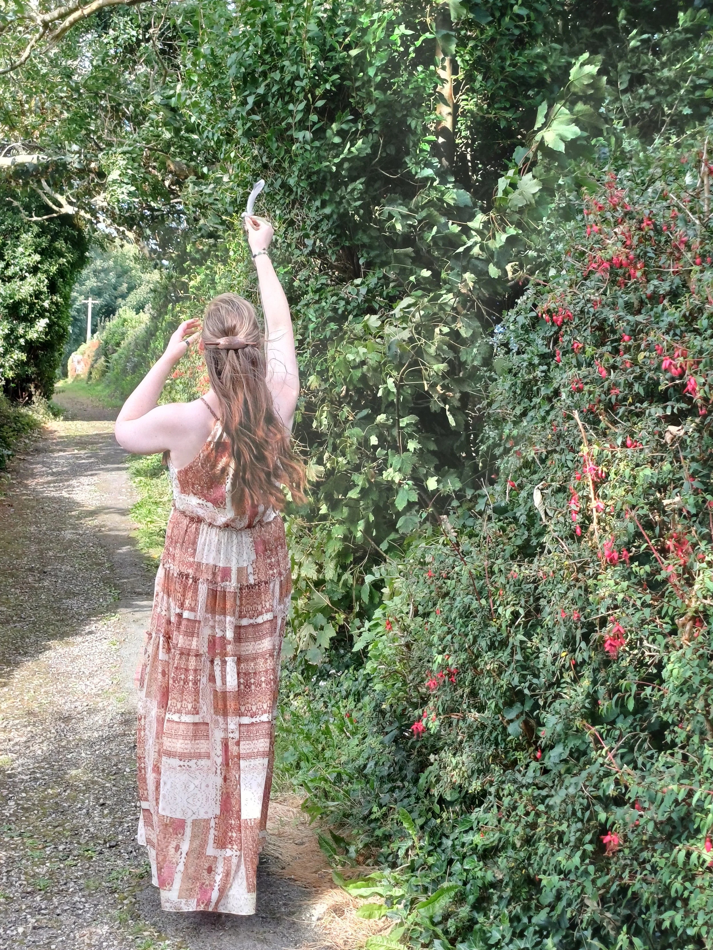 Christina Sayers standing on a nature path, raising a white feather to symbolise the spiritual signs, connection, and angelic presence that guide her work at Tides of Spirit.