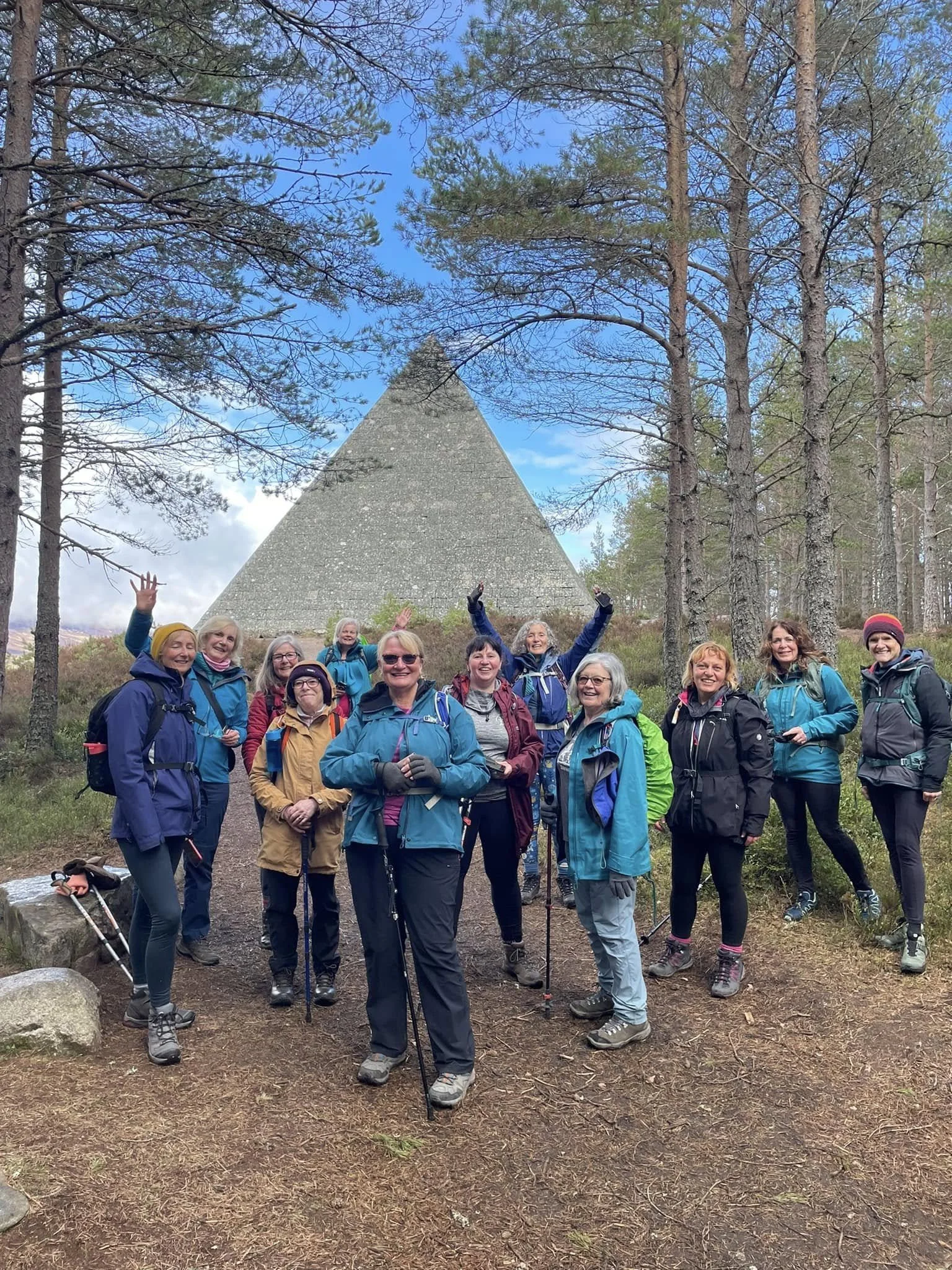 Group of eleven women hikers in outdoor gear posing in front of a large stone pyramid-shaped structure in a forested area with blue sky and clouds overhead.