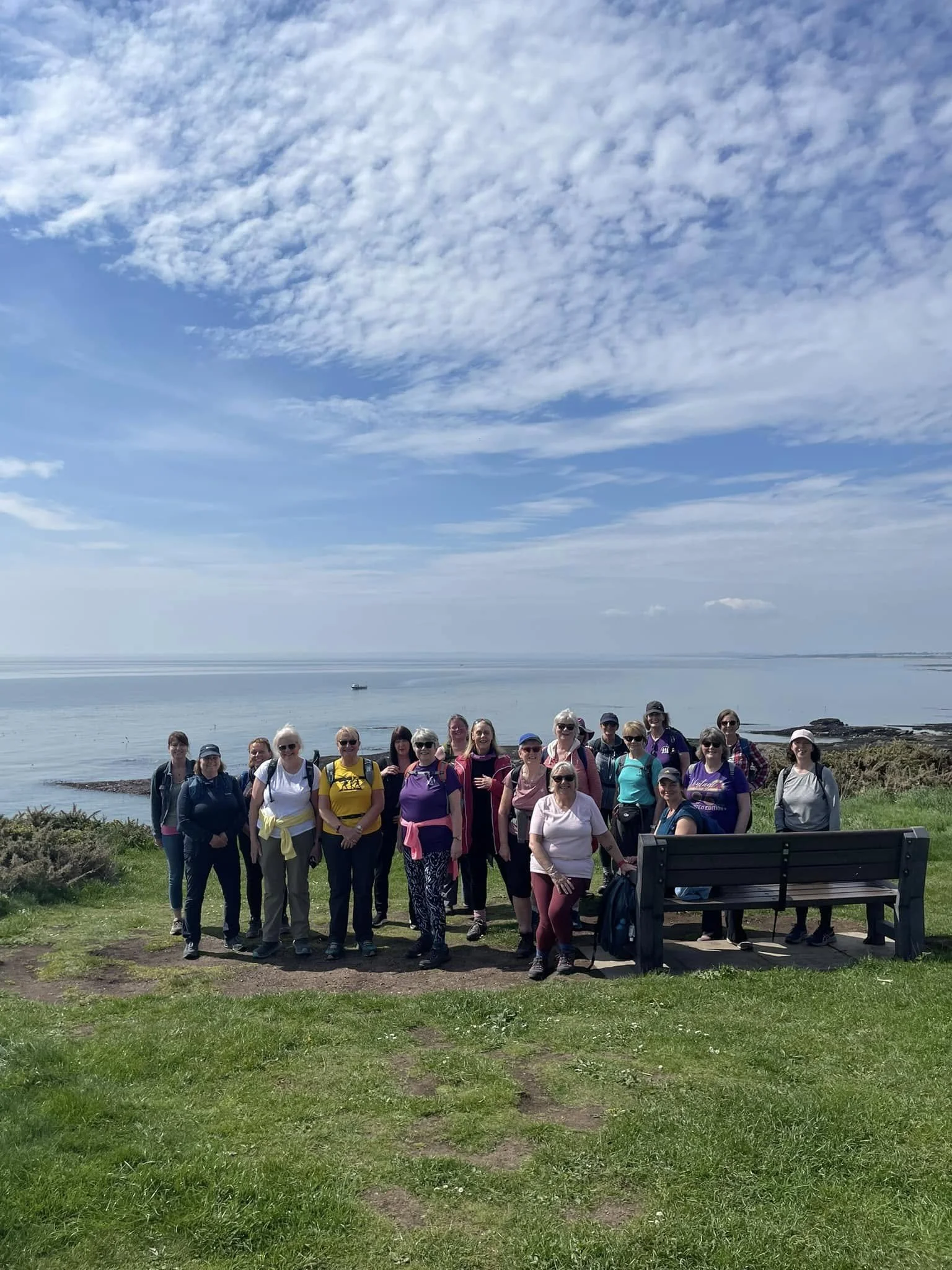 A group of people standing on a grassy area near the water with a cloudy sky in the background.