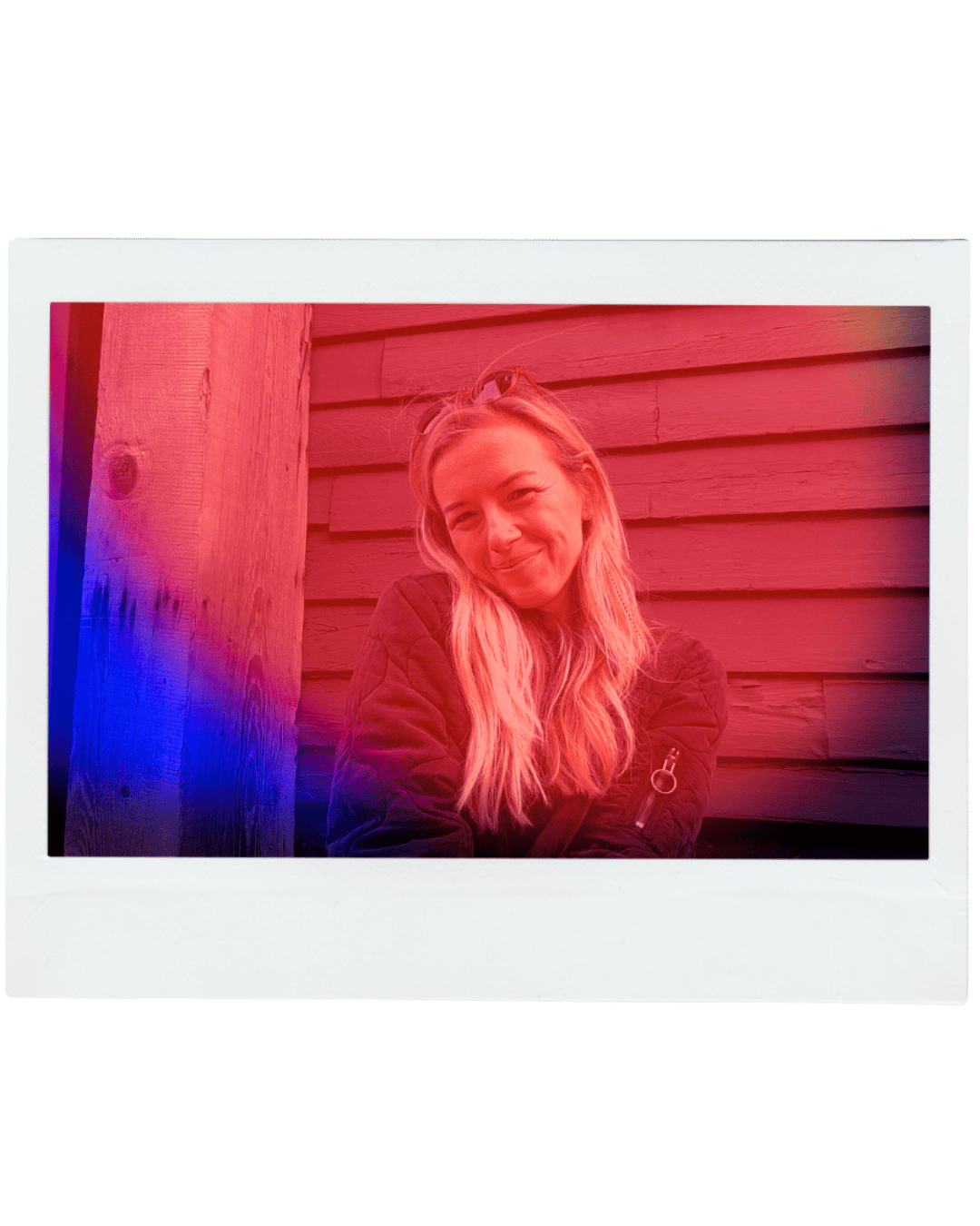 Photo of a smiling woman with long blonde hair, wearing a black jacket, sitting against a wooden wall with sunlight illuminating her face.