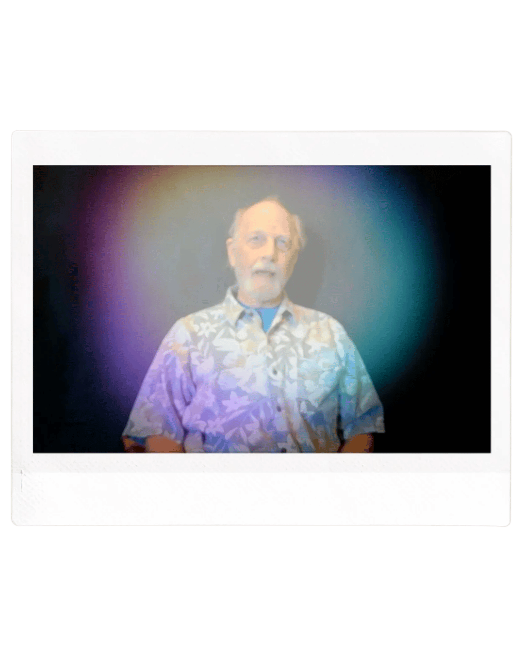 Older man with glasses and a beard wearing a floral Hawaiian shirt, standing against a black background with colorful light effects.