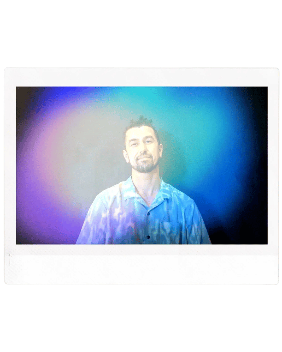 A man with dark hair and a beard wearing a light blue button-up shirt, looking at the camera, with a colorful gradient background.