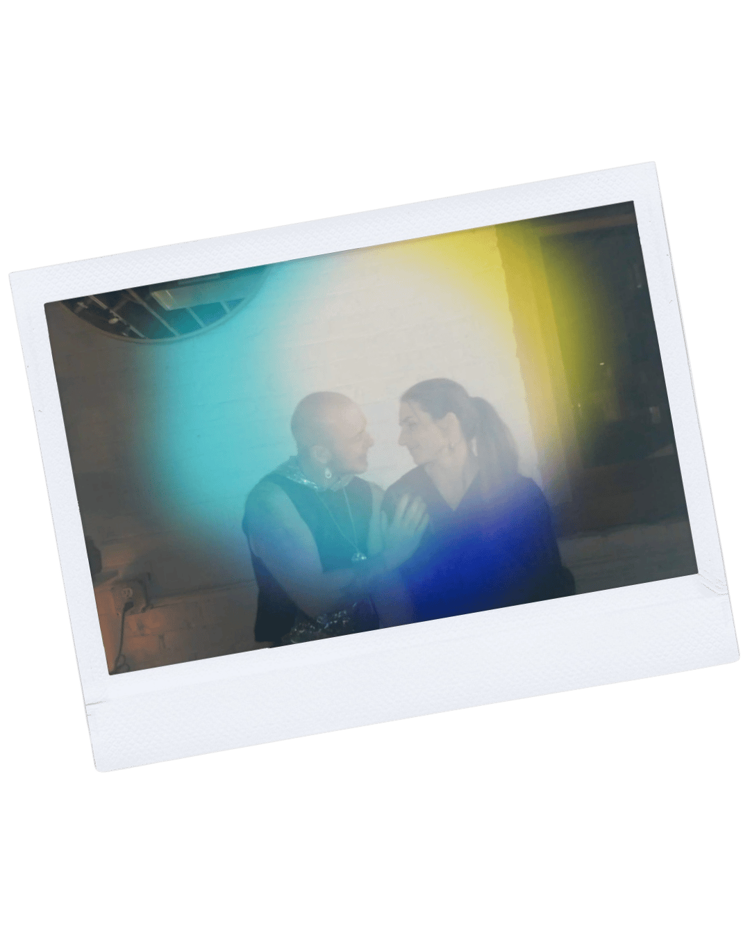 Two women engaging in conversation, with one touching the other's arm, in front of a white brick wall, with a rainbow-like lens flare.