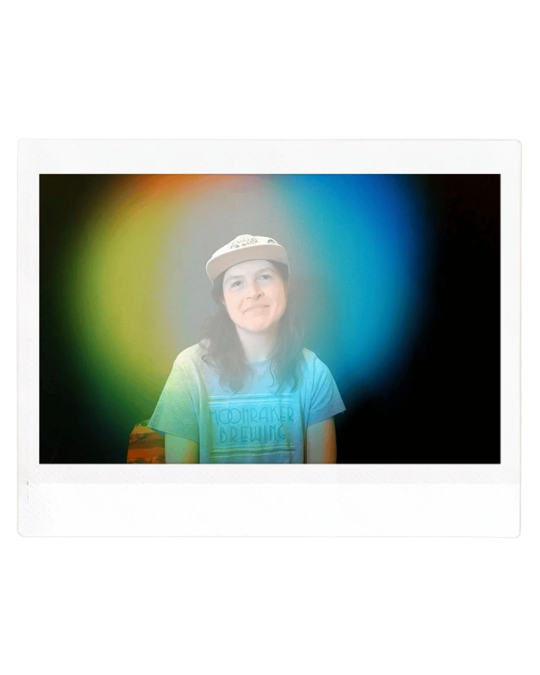 Person with long hair, wearing a baseball cap and a gray T-shirt with a logo, sitting in front of a dark background with a rainbow-colored glow around them.