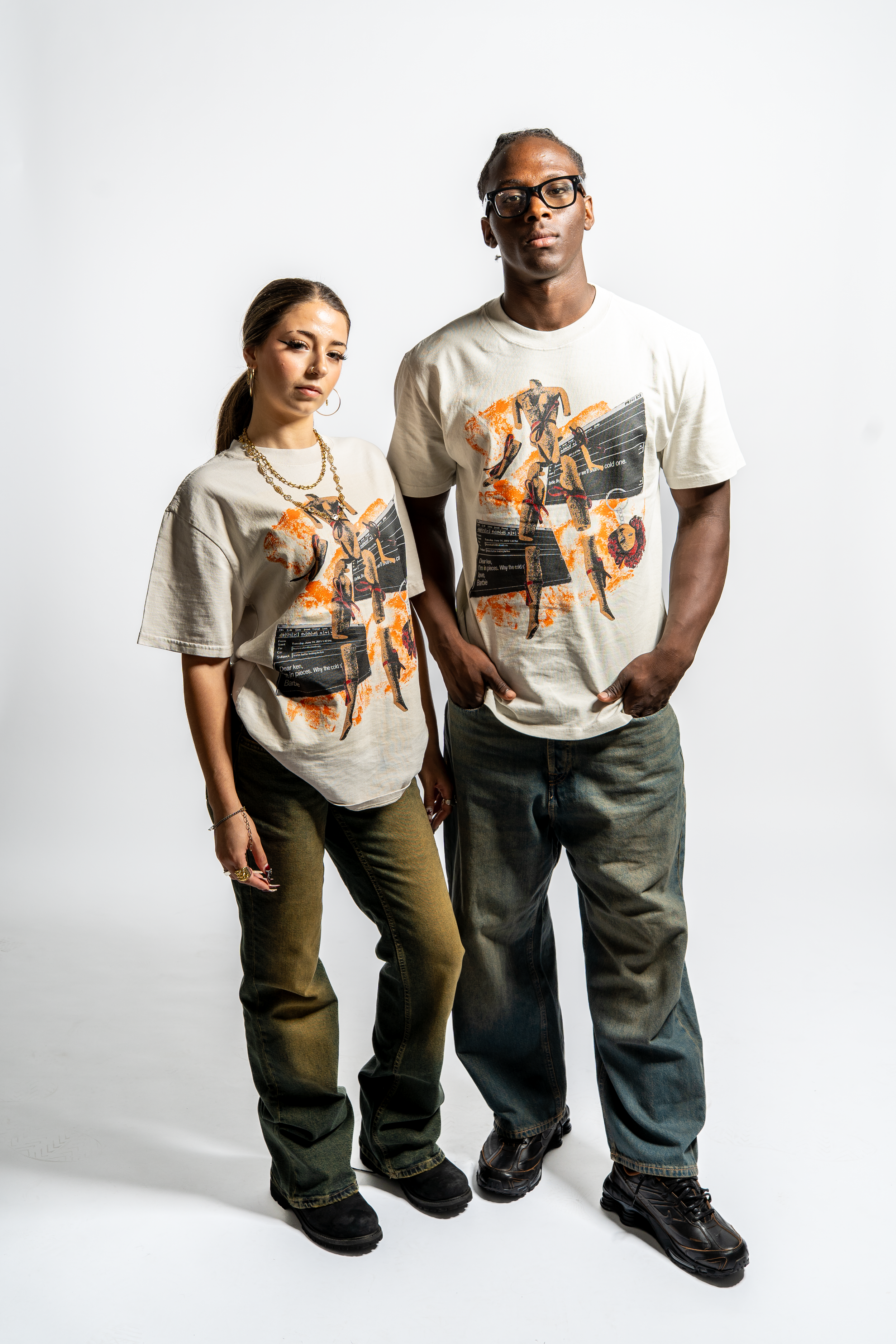 A man and woman standing against a plain white background, both wearing matching graphic t-shirts, dark jeans, and black shoes.
