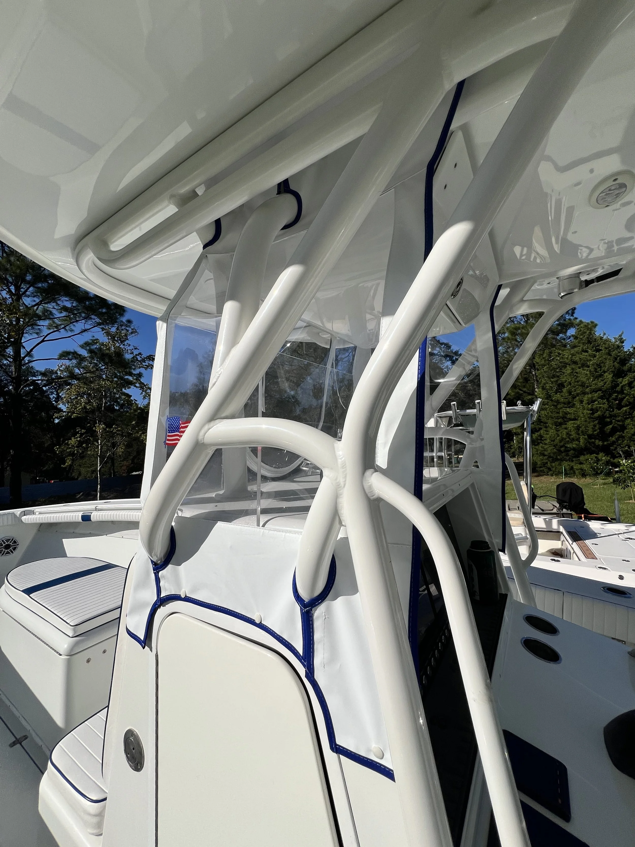 Close-up view of a white boat's helm and seating area, with a black cup holder, canopy support structures, and a clear windshield. Trees and blue sky are visible in the background.