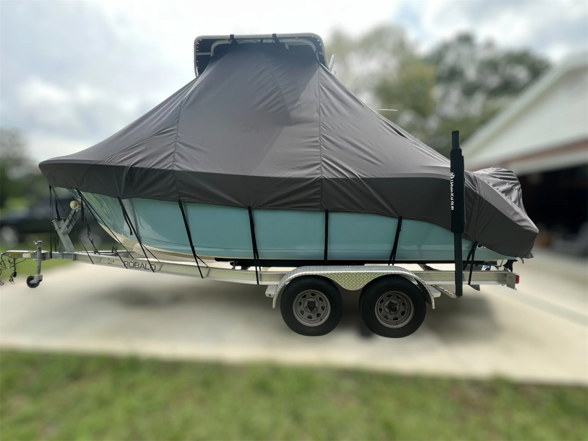 A boat on a trailer behind a vehicle, covered with a black boat cover, parked on a driveway with a house and trees in the background.