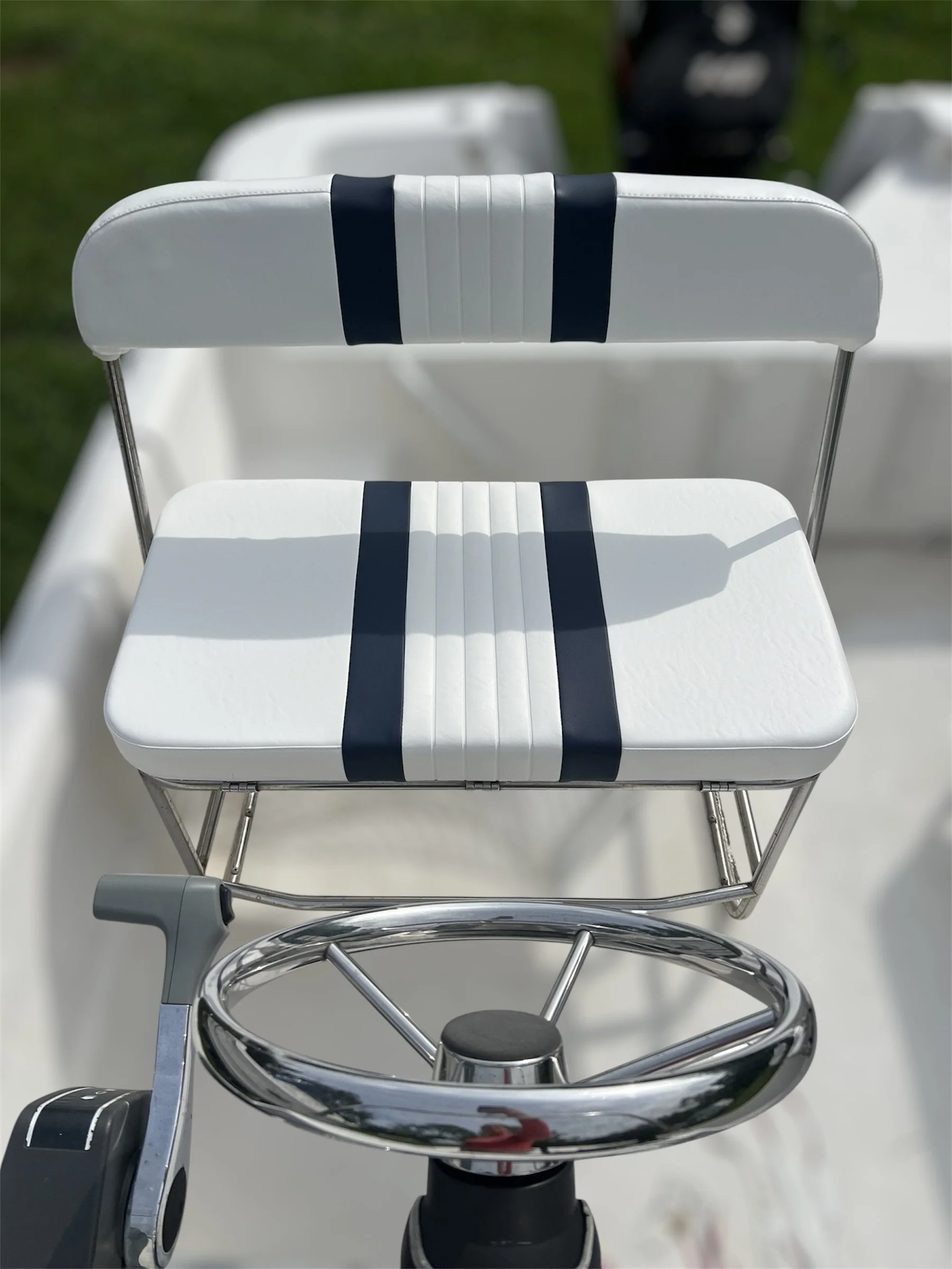 Close-up of a white golf cart with a padded seat and backrest, both with navy blue stripes, and a steering wheel in the foreground. Background shows a grassy area.
