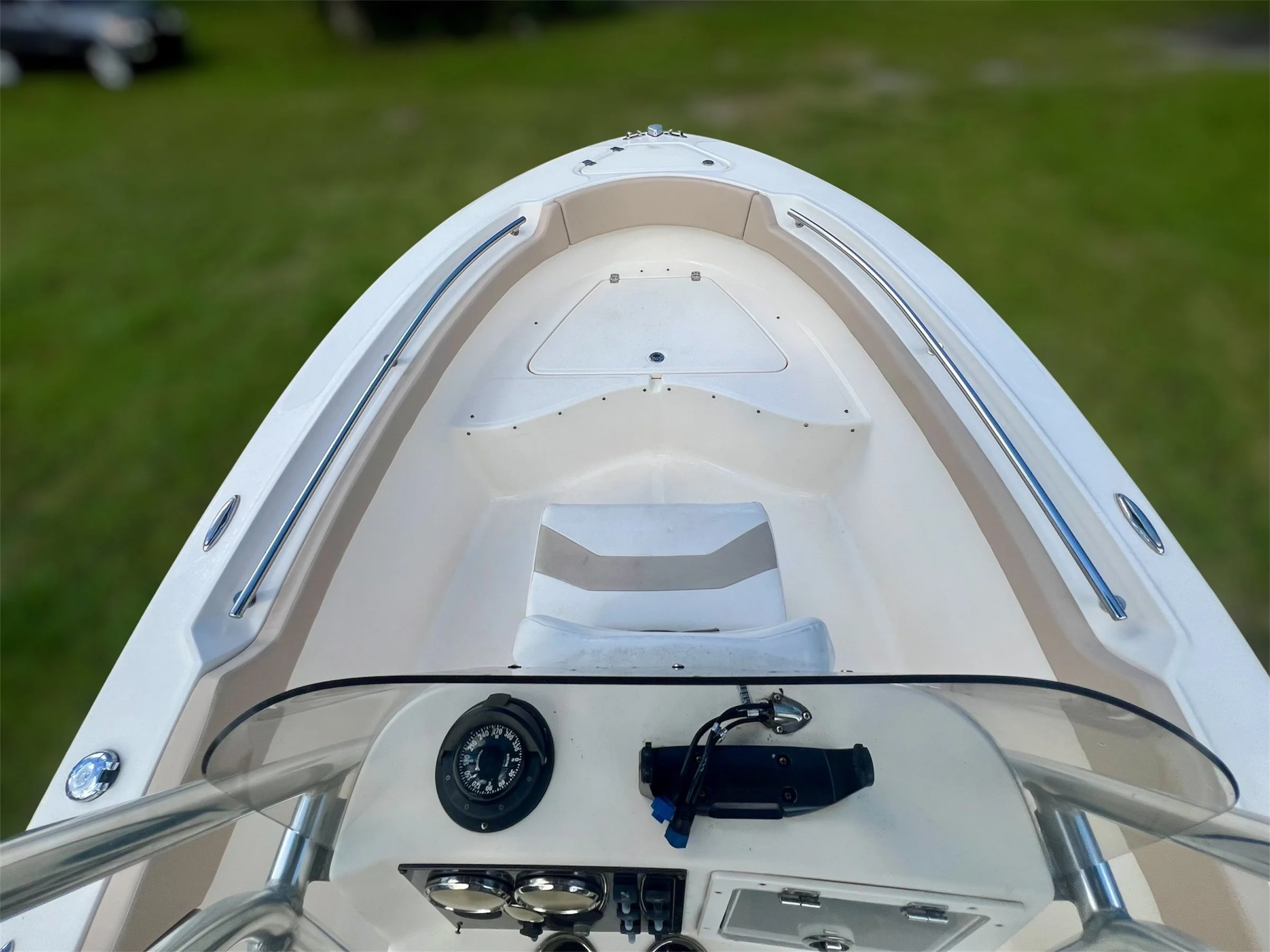 Top view of a small white motorboat with a cushioned seat at the bow, a steering console with a compass, controls, and a clear windshield, on a grassy area.