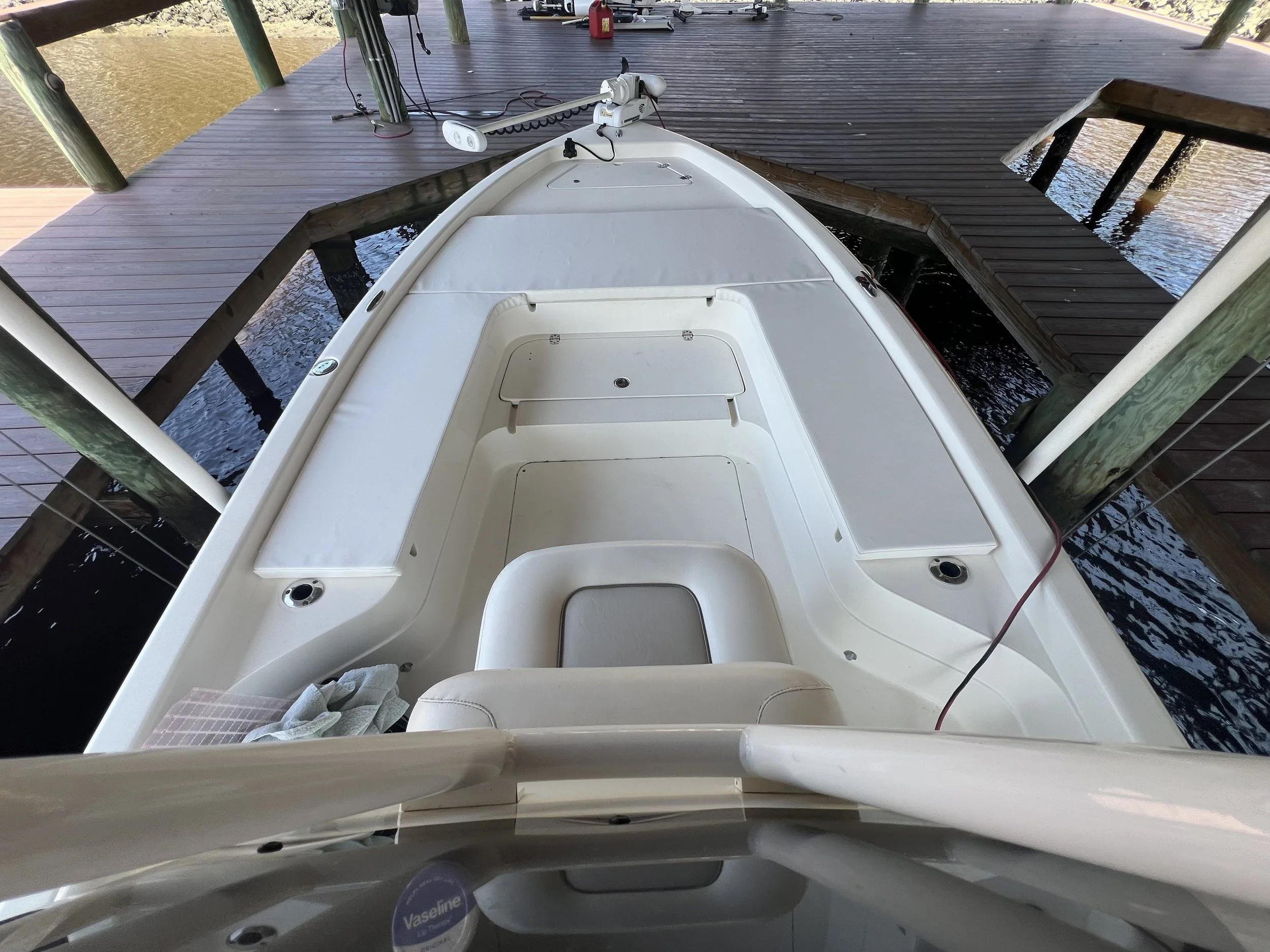 View from above of the front of a white boat docked at a wooden pier, showing the boat's deck with seating areas and compartments, docked at a calm water marina.