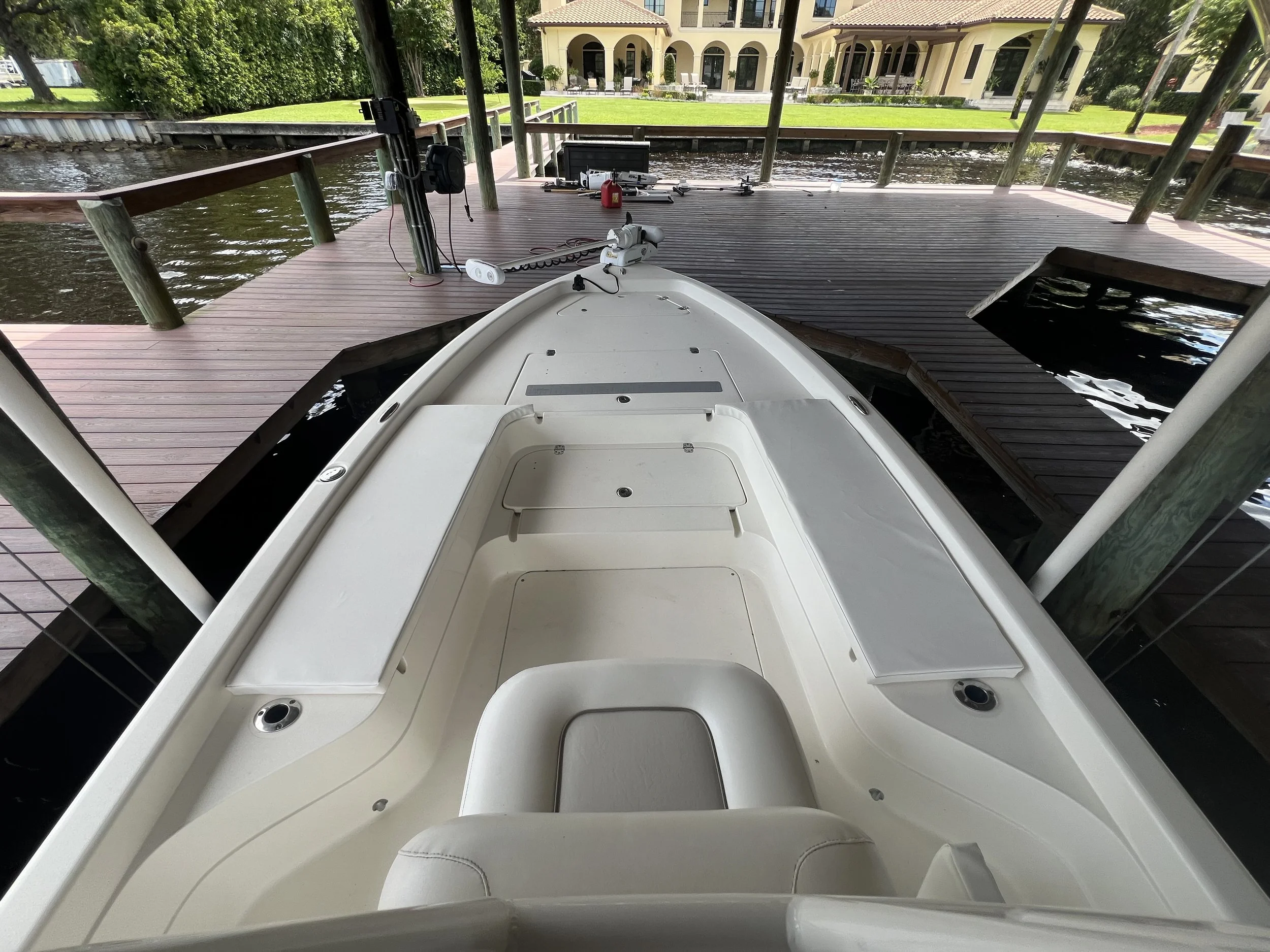 View from the front of a white boat docked at a wooden pier. The boat has seating and storage compartments. The pier extends into the water with residential houses and lush greenery in the background.