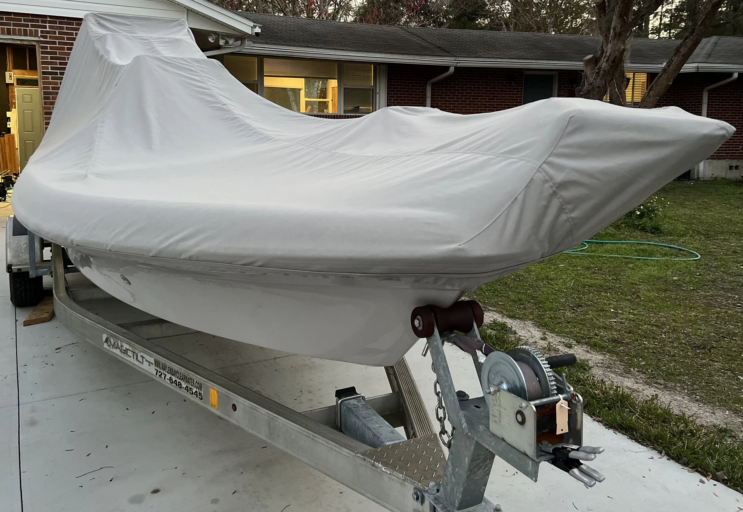 A boat covered with a protective grey cover on a trailer parked outside on a driveway next to a grassy yard, with a house in the background.