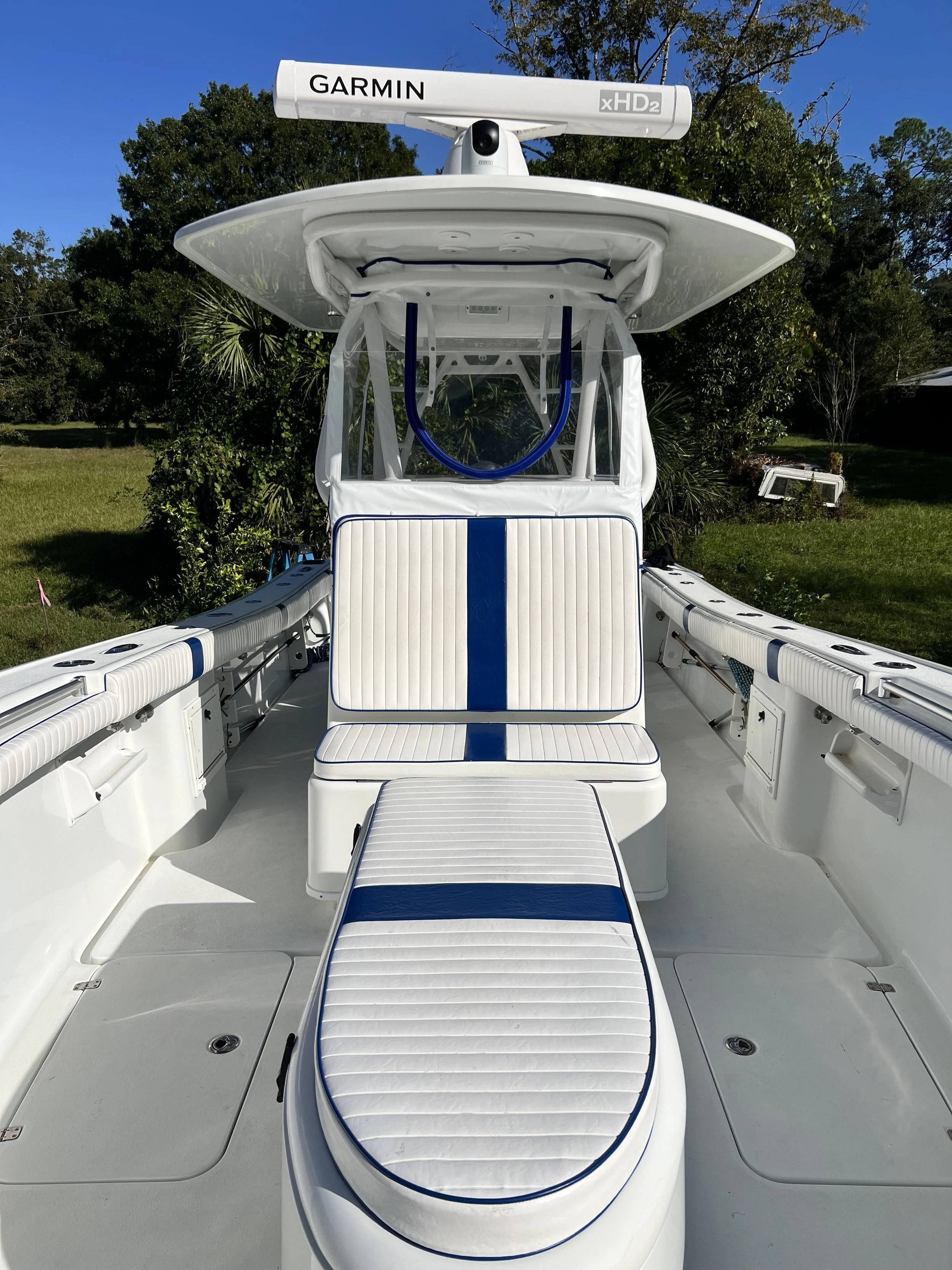 A white fishing boat with blue accents is parked outdoors on grass under a blue sky, featuring a Garmin xHD2 radar on top, a central cushioned seat, and side storage compartments.