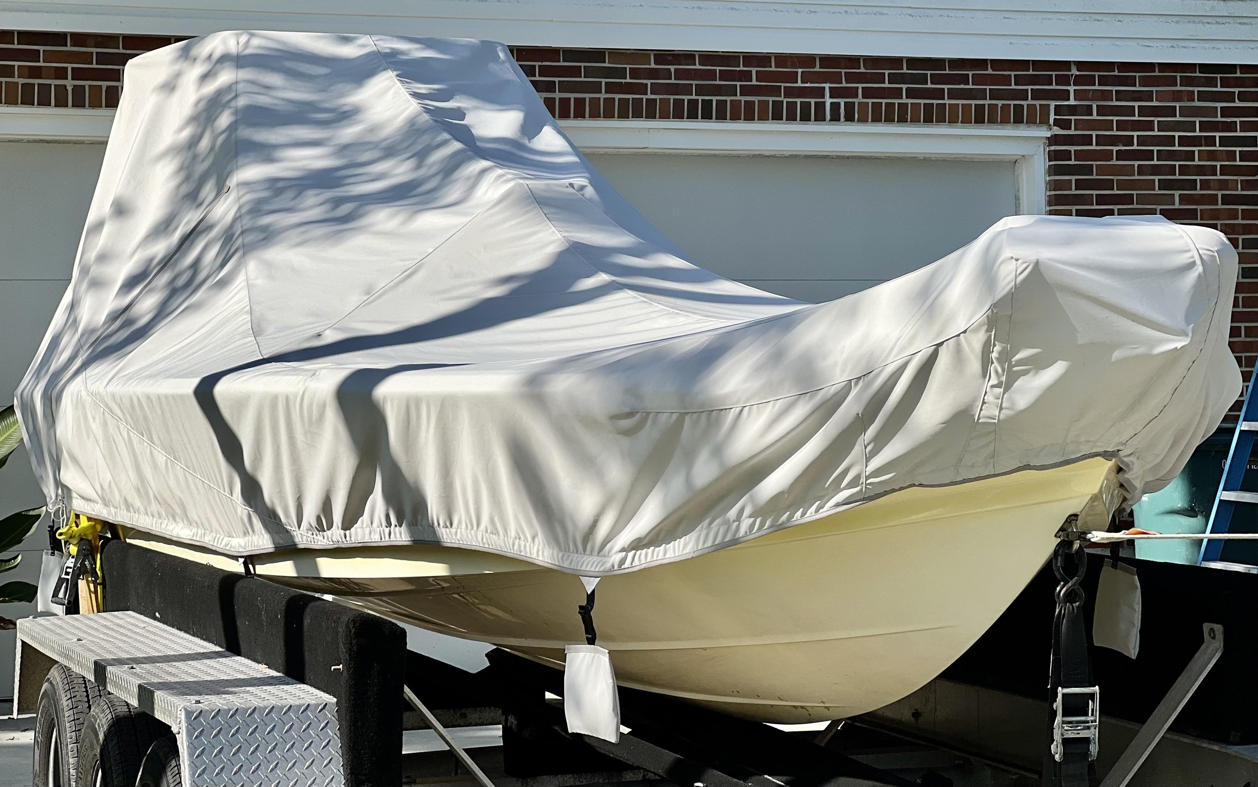 A boat on a trailer covered with a white protective cover, parked in front of a brick house.