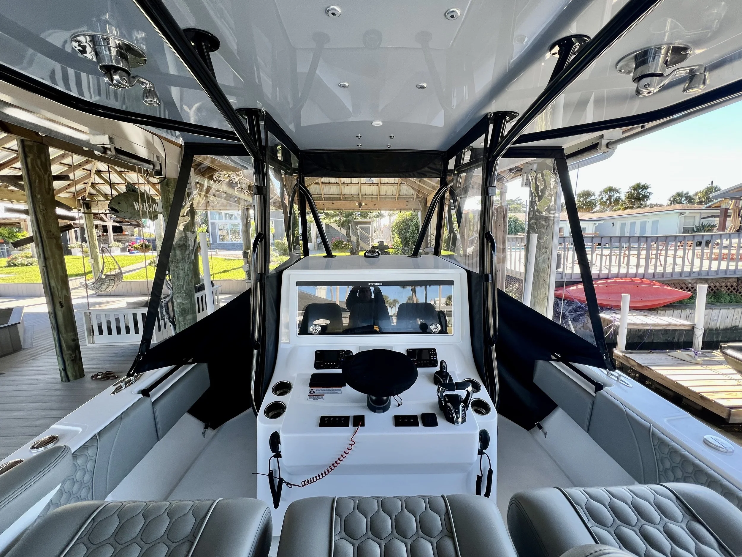 View of a boat's cockpit with a steering wheel, throttle controls, and a computer screen, surrounded by seats and windows showing an outdoor dock area.