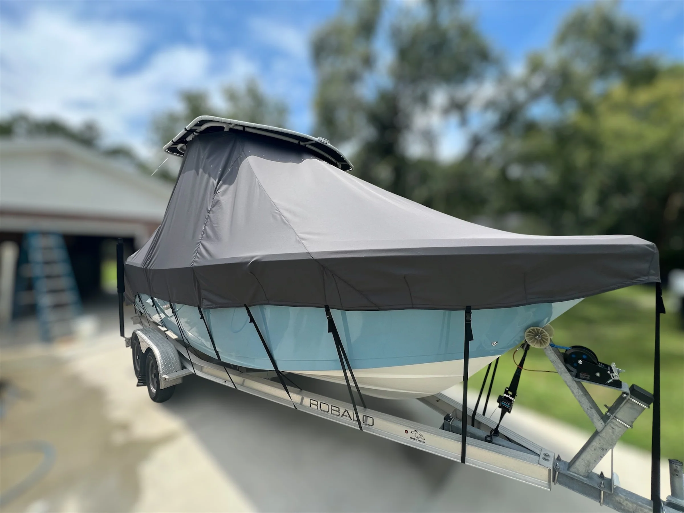 A small boat with a canopy cover is on a trailer parked outdoors, with a blurred background of trees and a house.