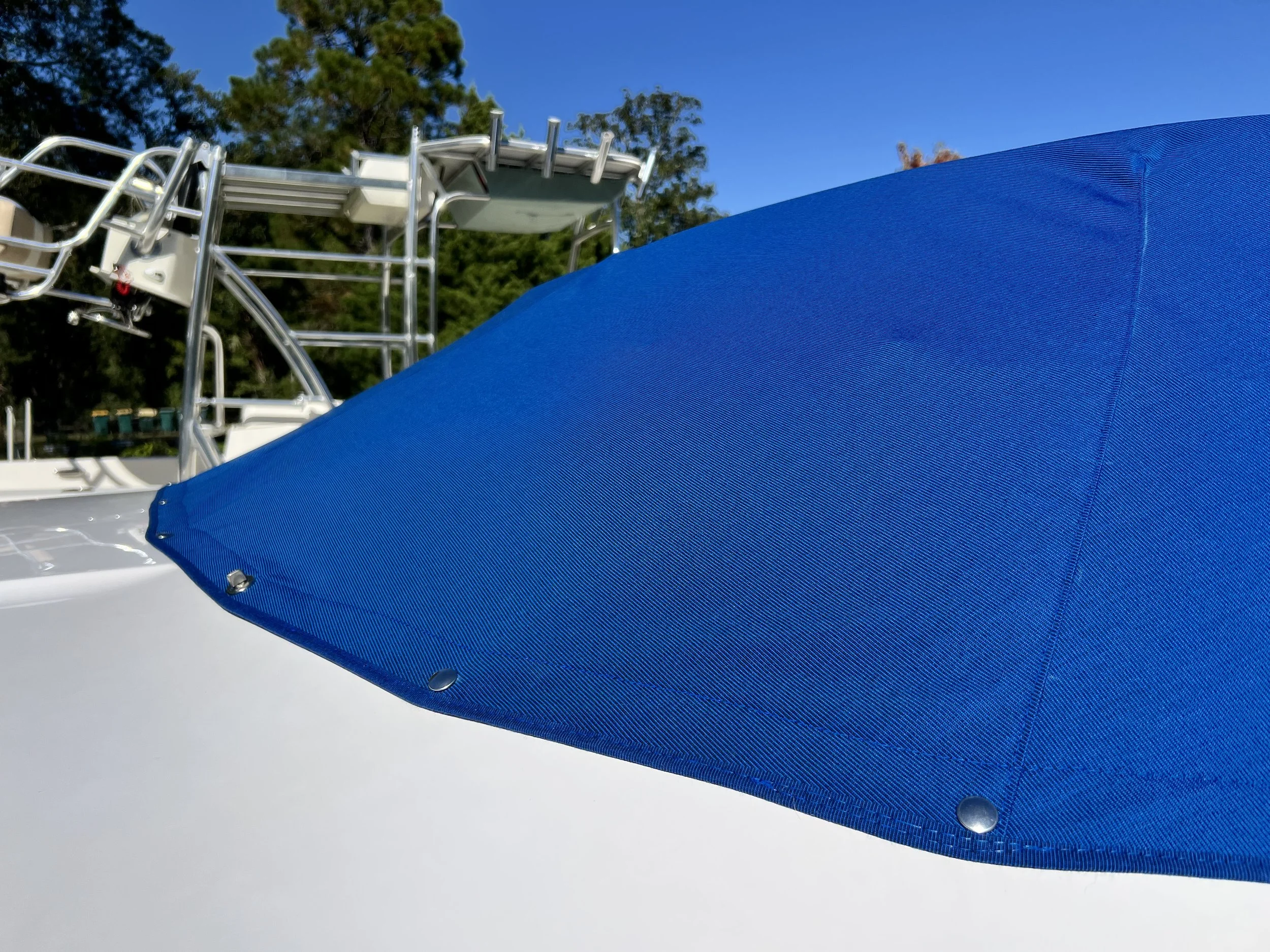 Close-up of a blue boat cover with metal rivets, with a boat's metal framework and trees in the background under a clear blue sky.